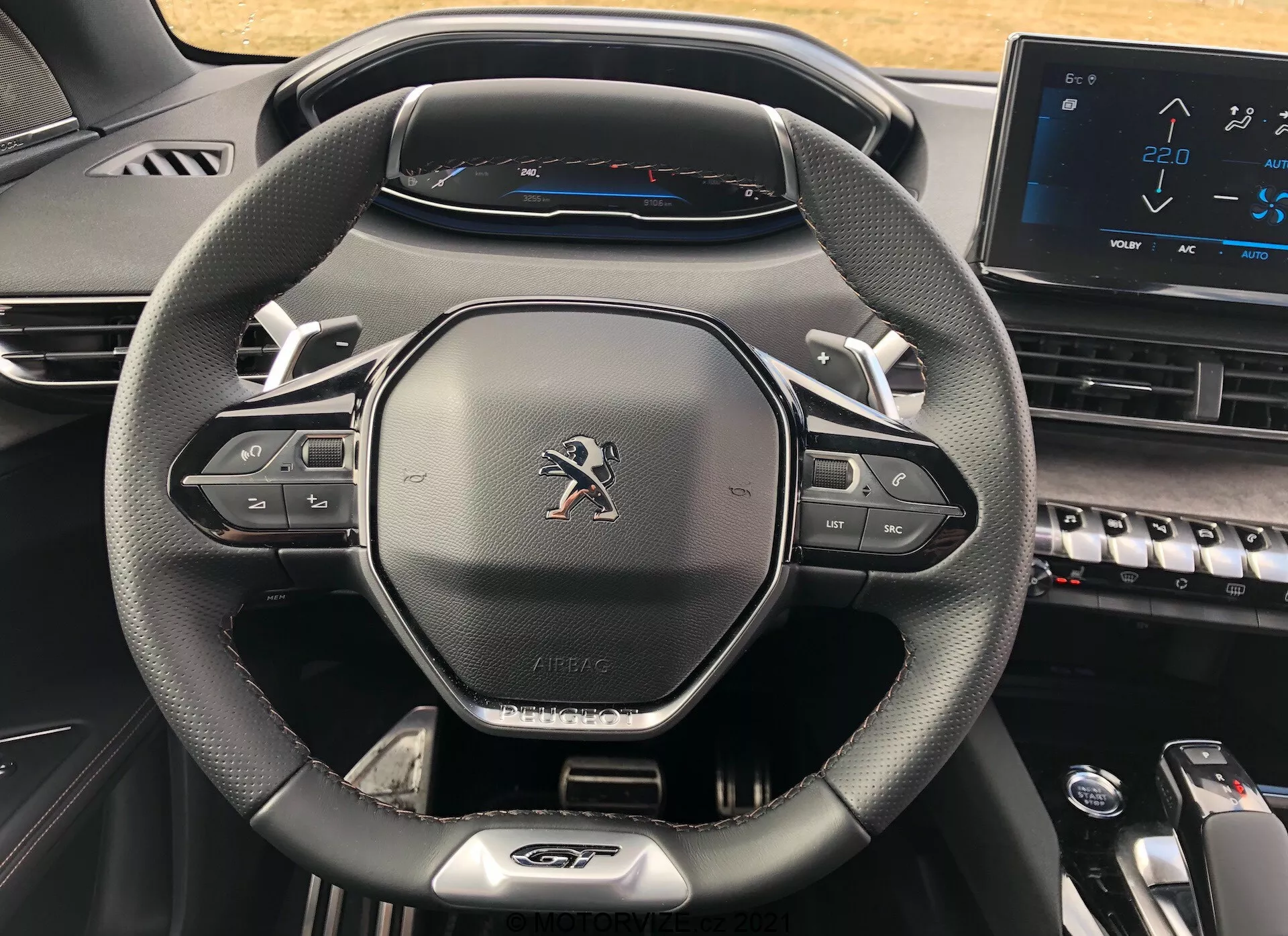 Zbliżenie kokpitu kierowcy w modelu Peugeot 5008 z 2021 roku, z naciskiem na nowoczesną, trójramienną kierownicę o teksturze skórzanej z przyciskami sterowania multimediami i tempomatem, emblemat Peugeot, etykietę 'AIRBAG', w pełni cyfrowy zestaw wskaźników wyświetlający prędkościomierz z maksymalną prędkością 220 km/h oraz część konsoli centralnej z klawiszami w stylu fortepianu i elektronicznym ekranem dotykowym, co przedstawia nowoczesny i technologicznie zaawansowany design.