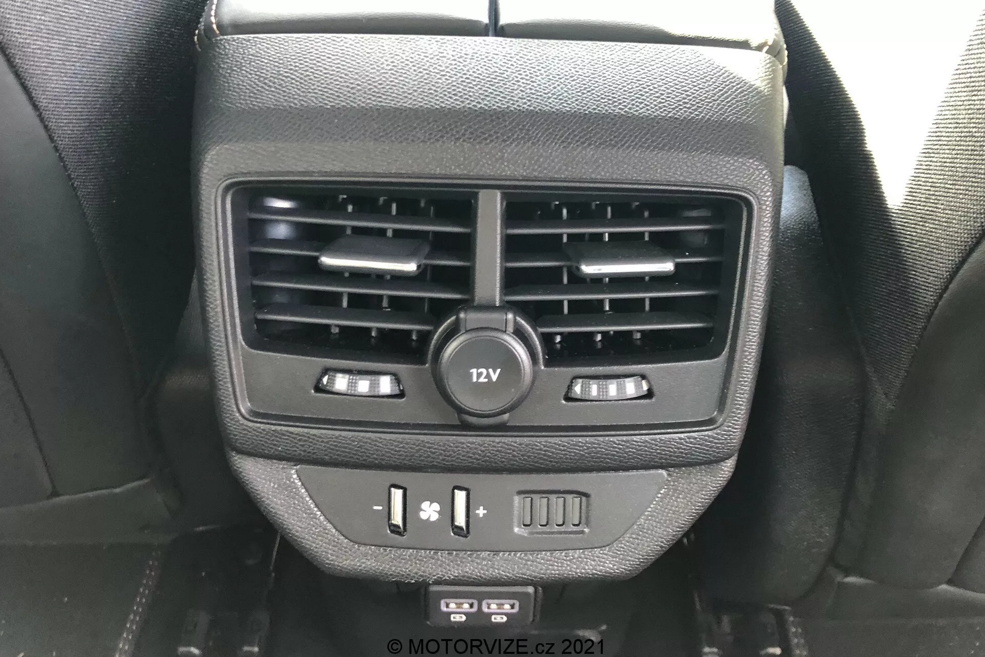 Zbliżony widok na funkcje dla pasażerów tylnych na tylnej części konsoli centralnej w samochodzie Peugeot 5008 2021. Obraz pokazuje podwójne wyloty powietrza, gniazdo zasilania 12V, sterowanie podgrzewaniem siedzeń, porty USB i pusty przycisk, uchwycony z perspektywy pasażera tylnego siedzenia.