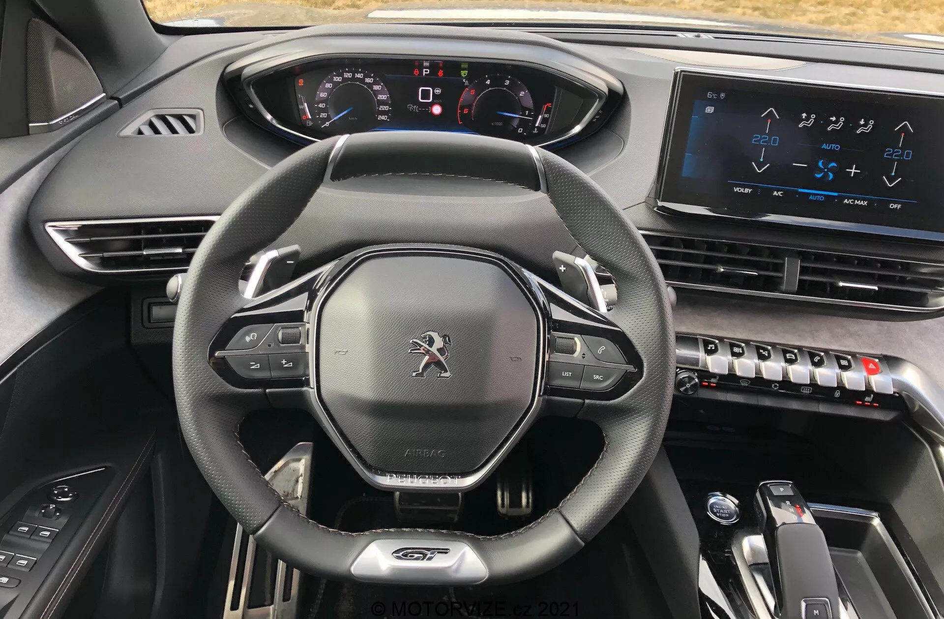 Widok wnętrza Peugeot 5008 2021 z perspektywy kierowcy, podkreślający skórzaną kierownicę z płaskim dnem z zamontowanymi kontrolerami, w pełni cyfrowy zestaw wskaźników z podświetlanymi wskaźnikami, duży ekran dotykowy systemu infotainment na konsoli centralnej, eleganckie sterowanie klimatyzacją, elektryczny wskaźnik zmiany biegów dla automatycznej skrzyni biegów oraz nowoczesne, wysokiej jakości materiały ciemne, które wywołują nowoczesną, high-tech i premium estetykę.