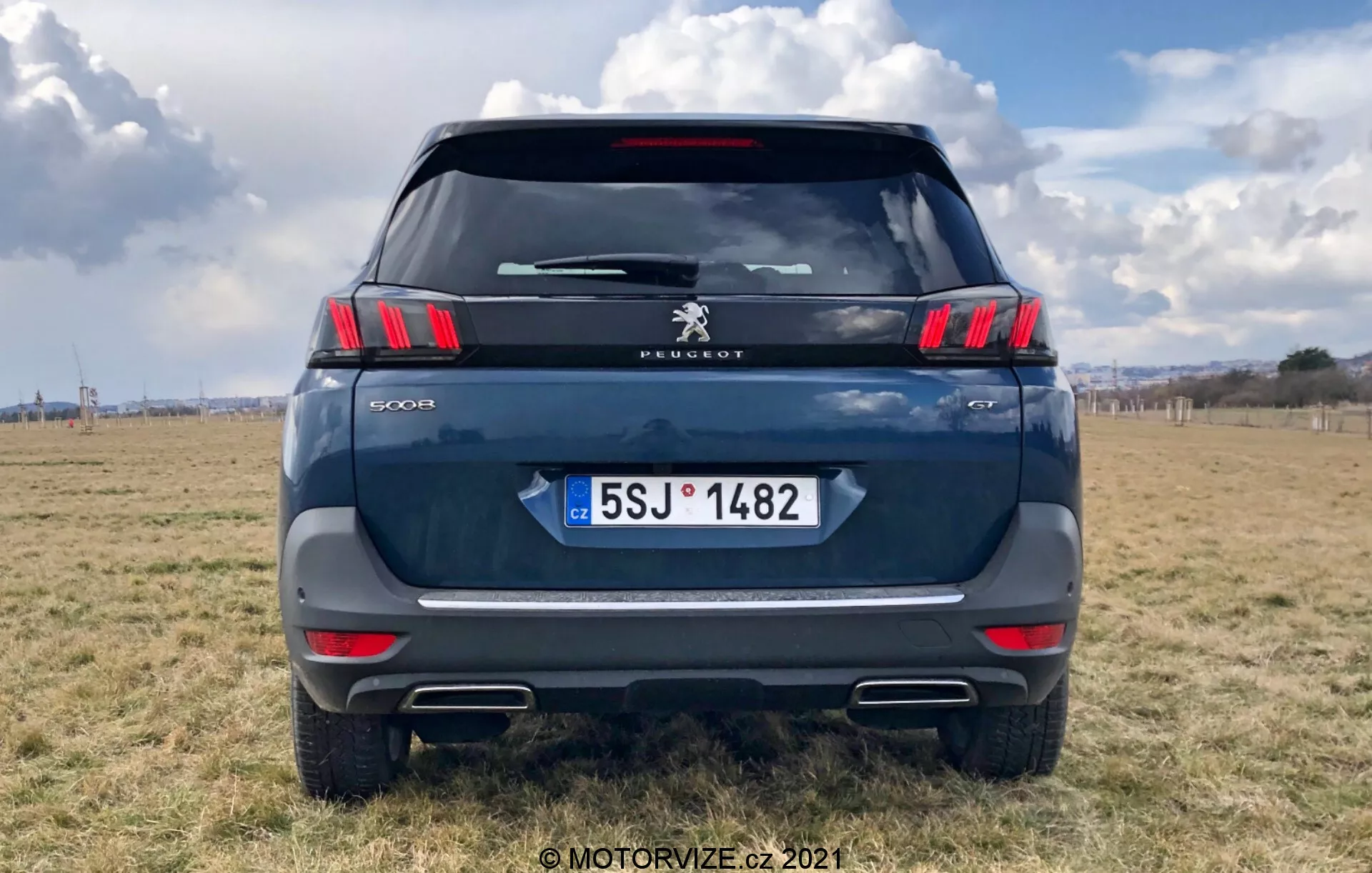 Widok z tyłu nowoczesnego SUV-a Peugeot 5008, podkreślający eleganckie poziome tylne światła z charakterystycznymi segmentami LED w kształcie pazurów, logo Peugeot i napis modelu '5008'. Kolor ciemnoniebieski lub szary, zaparkowany na zewnętrznym terenie z częściowo zachmurzonym niebem. Widoczne elementy to tylna szyba, wycieraczka tylna, wysoko montowana lampa stopu, uchwyt klapy tylnej i tablica rejestracyjna Republiki Czeskiej z 'CZ'. Pod paskiem odblaskowym z czujnikami parkowania i wycięciami imitującymi końcówki wydechów widoczne są wyraźnie. Ujęcie centralne z samochodem wypełniającym większość kadru.
