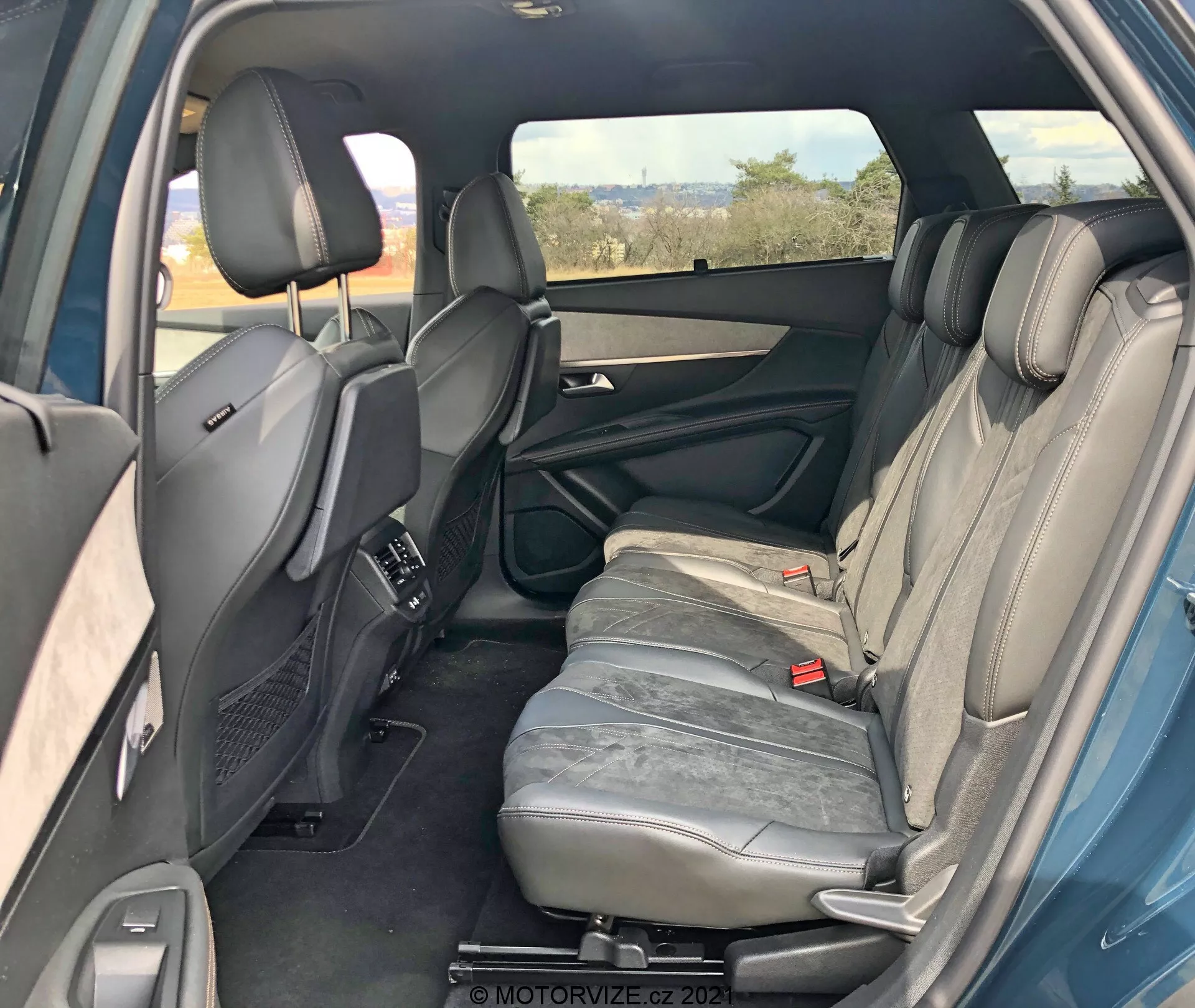 Wnętrze Peugeot 5008 2021 z perspektywy prawego tylnego pasażera, ukazujące tylny przedział pasażerski. Prawe tylne drzwi są otwarte, ukazując cechy panelu drzwiowego, takie jak uchwyt, schowki i głośnik. Widoczne są trzy siedzenia tylne obite szarą skórą z zagłówkami. Środkowe oparcie siedzenia jest w pozycji pionowej. Widoczne są również tylne pasy bezpieczeństwa oraz przednie siedzenia z regulowanymi zagłówkami i konsolą centralną. Tylne kratki wentylacyjne na konsoli środkowej, przyciemniana prawa szyba, przebłyski lewych tylnych drzwi wskazujące symetrię. Ogólnie dobrze utrzymane wnętrze bez widocznych cech technologicznych.