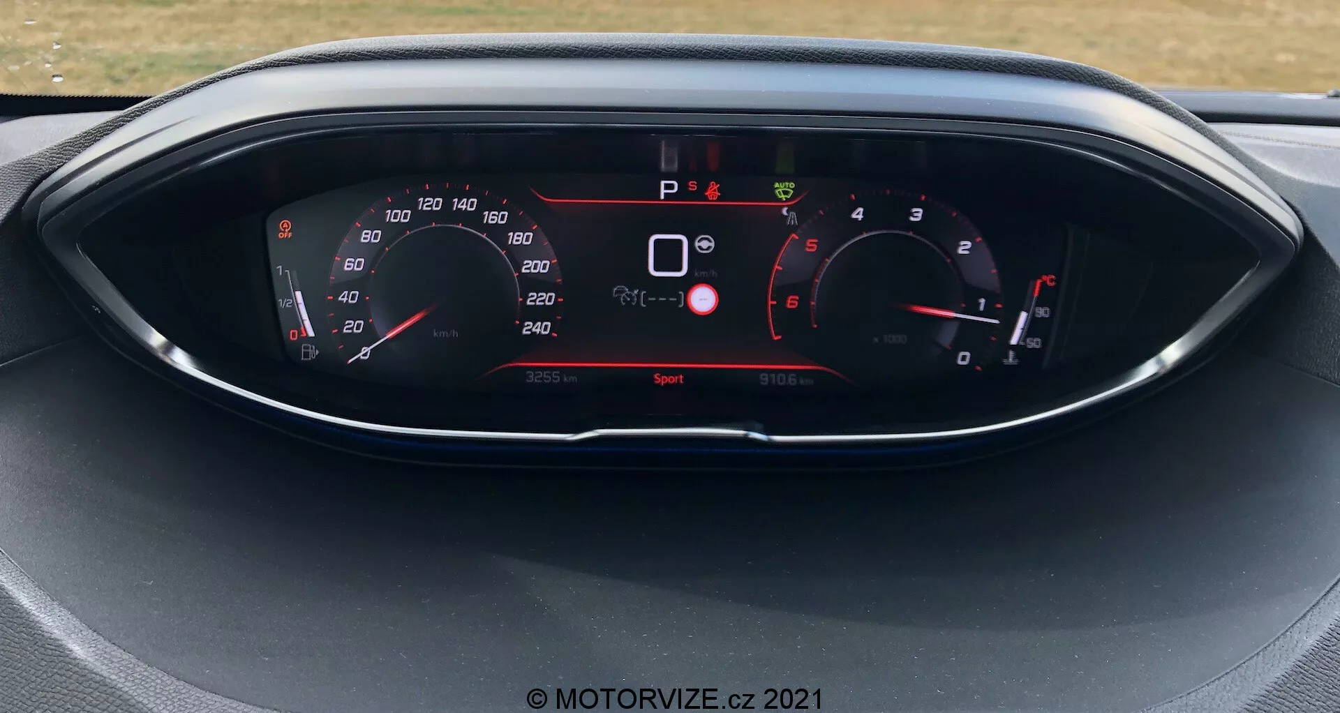 Zbliżenie na panel instrumentów w Peugeot 5008 z 2021 roku, z cyfrowym wyświetlaczem i wskaźnikami w stylu analogowym. Wyświetlacz pokazuje prędkościomierz, obrotomierz, wybór biegu oraz różne wskaźniki pojazdu, takie jak ostrzeżenia bezpieczeństwa i tryb jazdy. Samochód jest unieruchomiony, a aktywny jest tryb jazdy 'Sport'.