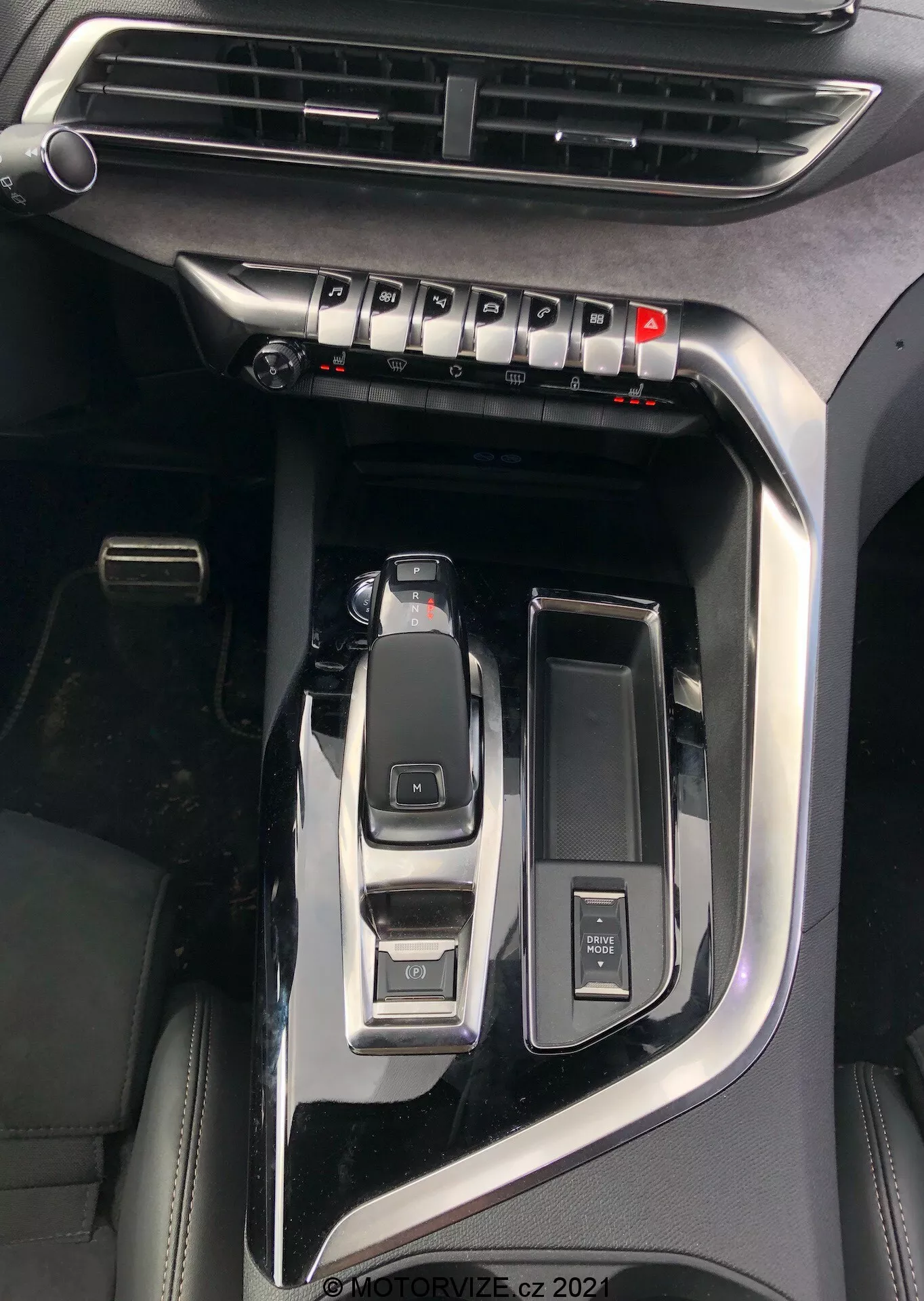 Zbliżenie konsoli środkowej Peugeot 5008 2021 podkreślające kluczowe cechy, takie jak wyraźna dźwignia zmiany biegów automatycznych w pozycji parkowania, selektor trybów jazdy do trybów eco, sport, comfort, przycisk start/stop do bezkluczykowego uruchamiania, pokrętło sterowania multimediami, otwarty uchwyt na kubek, widoczny nawiew klimatyzacji oraz wykończenie w czarnym fortepianie z metalicznymi akcentami, co wskazuje na model premium, znak wodny 'MOTORVIZE.cz 2022'.