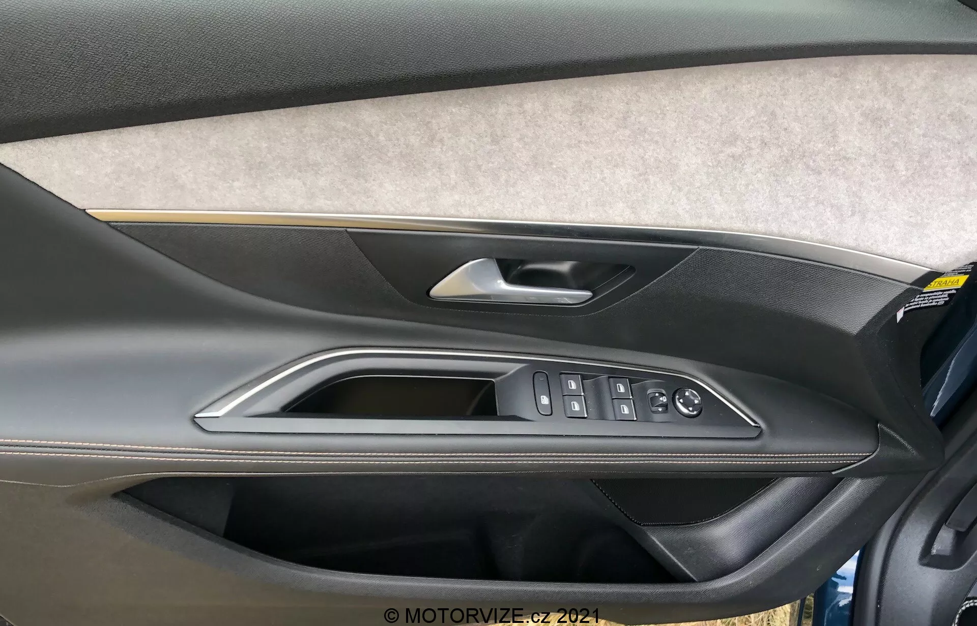 Zbliżenie na wewnętrzny panel drzwiowy Peugeot 5008 2021 z sekcją wykończoną filcem lub tkaniną, dwukolorową konstrukcją z formowanego plastiku z ciemniejszą górną częścią i jaśniejszą dolną częścią, wysokiej jakości metalową lub chromowaną klamką drzwi, polerowaną listwą oraz ergonomicznymi przyciskami sterującymi do elektrycznych szyb, zamka drzwi i lusterek bocznych. Na dole panelu drzwiowego widoczna jest również kieszeń na przechowywanie.