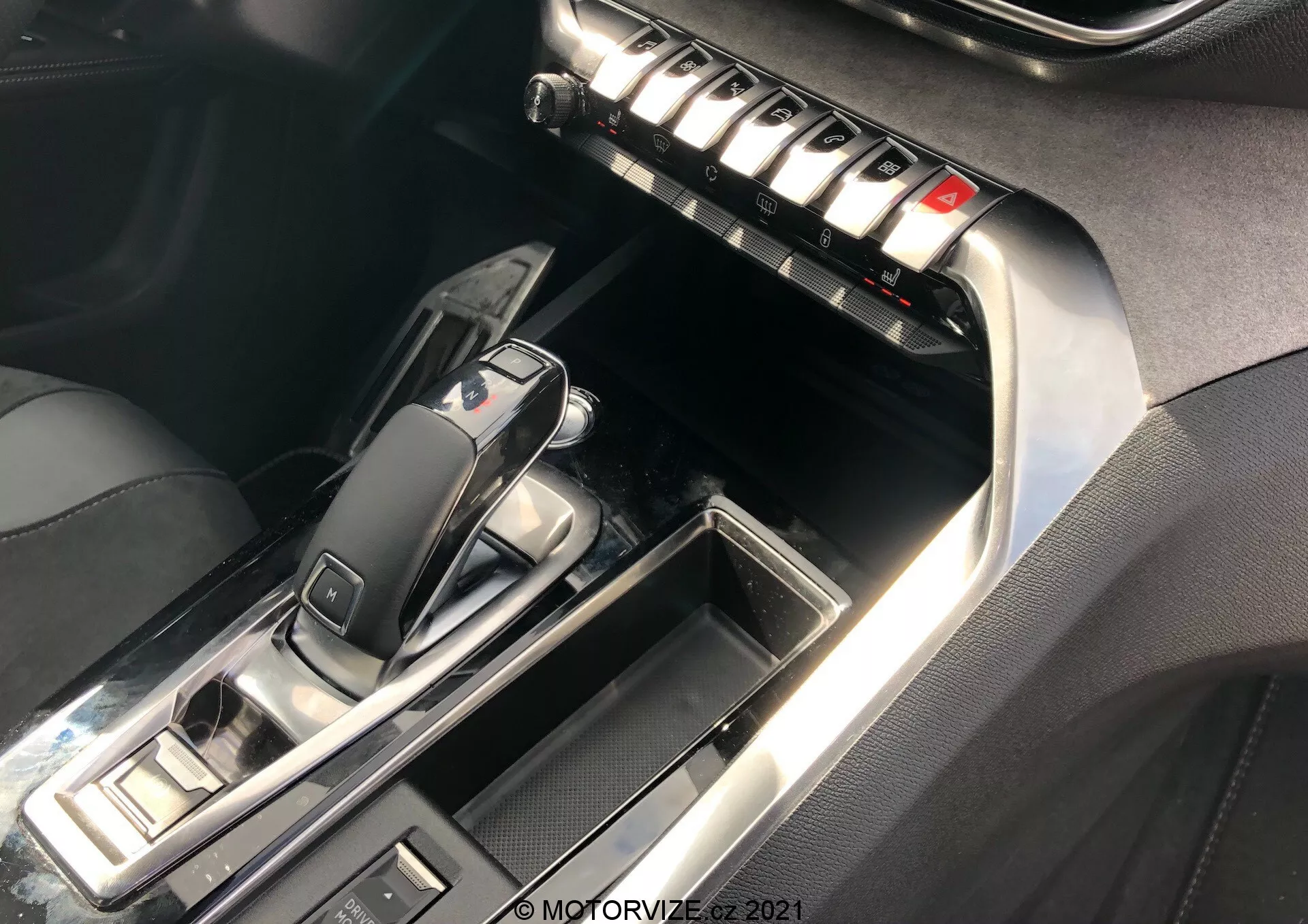 Zbliżenie konsoli środkowej i przedniego wnętrza Peugeot 5008 2021, skupiając się na obszarze automatycznej skrzyni biegów i środkowym panelu. Dźwignia zmiany biegów, w położeniu 'P', charakteryzuje się eleganckim designem z połyskującym czarnym plastikiem i metalicznymi wykończeniami. Otaczające przyciski sugerują tryby jazdy i sterowanie hamulcem postojowym. Panel środkowy obejmuje rząd metalicznych przycisków prawdopodobnie do sterowania klimatyzacją, odmrażaczami i ustawieniami siedzeń. Kontrastowe szwy na skórze lub ekoskórze i chromowane akcenty podkreślają luksusowy charakter wnętrza. Oświetlenie podkreśla refleksyjne powierzchnie i detale.
