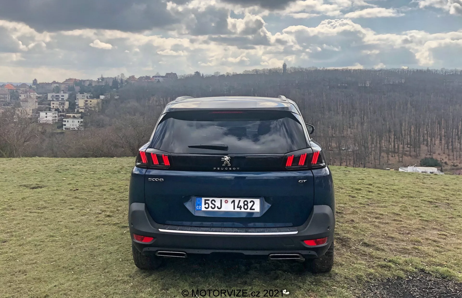 Bezpośredni widok tyłu SUV-a Peugeot 5008 2021 na podwyższonym trawiastym obszarze z panoramą miasta w tle, prezentujący tylne światła LED, czujniki parkowania zintegrowane z zderzakiem, możliwą kamerę cofania, tablicę rejestracyjną w stylu europejskim, podwójne końcówki wydechu, spojler dachowy i antenę w kształcie płetwy rekina; sceneria dzienna sugeruje częściowe zachmurzenie, sfotografowany przez MotorVize.cz w 2022 roku.