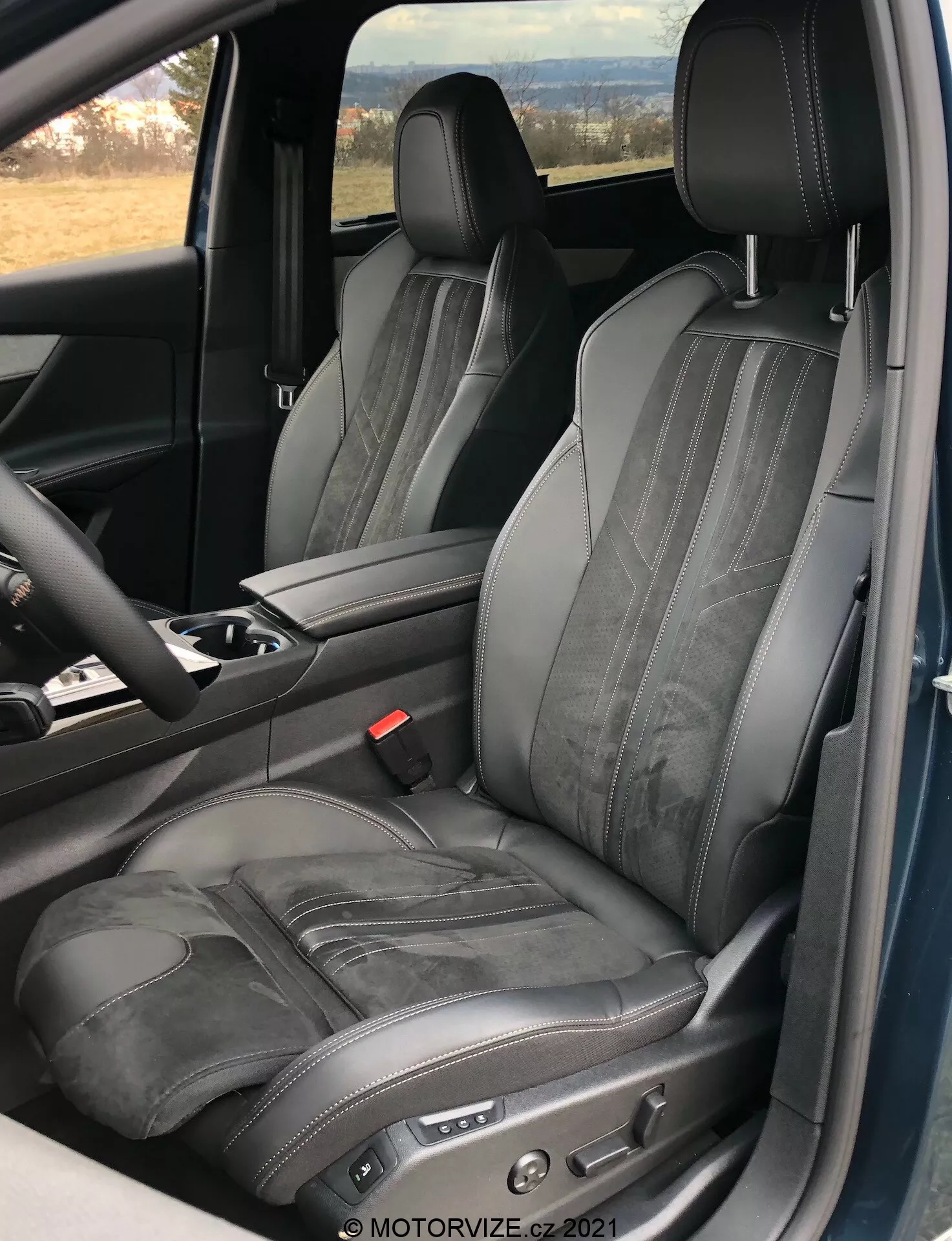 Widok wnętrza Peugeot 5008 2021 skupiony na fotelu kierowcy, pokazujący regulowany czarny skórzany fotel z kontrastowym przeszyciem i poziomym tłoczeniem, zrobiony z perspektywy otwartych drzwi, podkreślający luksusowy i ergonomiczny design z elektronicznymi przyciskami regulacji fotela oraz widoczną częścią konsoli centralnej i kierownicy.
