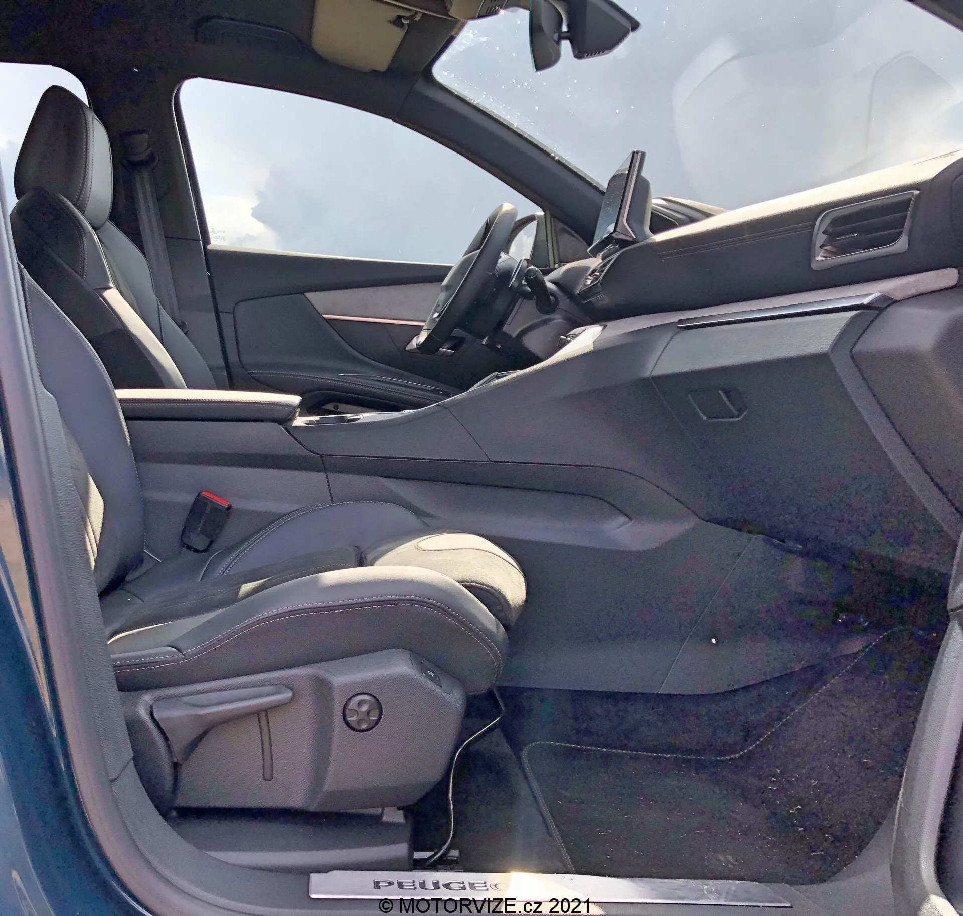 Widok wnętrza Peugeot 5008 z 2021 roku z miejsca kierowcy przy otwartych drzwiach. Fotel kierowcy jest kubełkowy, obszyty prawdopodobnie skórą lub materiałem skórzanym, z bocznymi podparciami i elektrycznymi elementami regulacyjnymi. Otwarta strona drzwi kierowcy pokazuje panel wewnętrzny z elektrycznymi kontrolkami okien, prawdopodobnie także lusterkami elektrycznymi i mechanizmami zamków drzwi. Deska rozdzielcza zawiera centralnie umieszczony ekran dotykowy infotainment z minimalistycznym designem. Widoczna górna część kierownicy sugeruje kierownicę obszytą skórą z zamontowanymi kontrolkami. Wnętrze ma dwutonową kolorystykę, z ciemniejszymi górnymi powierzchniami i jaśniejszymi dolnymi powierzchniami.