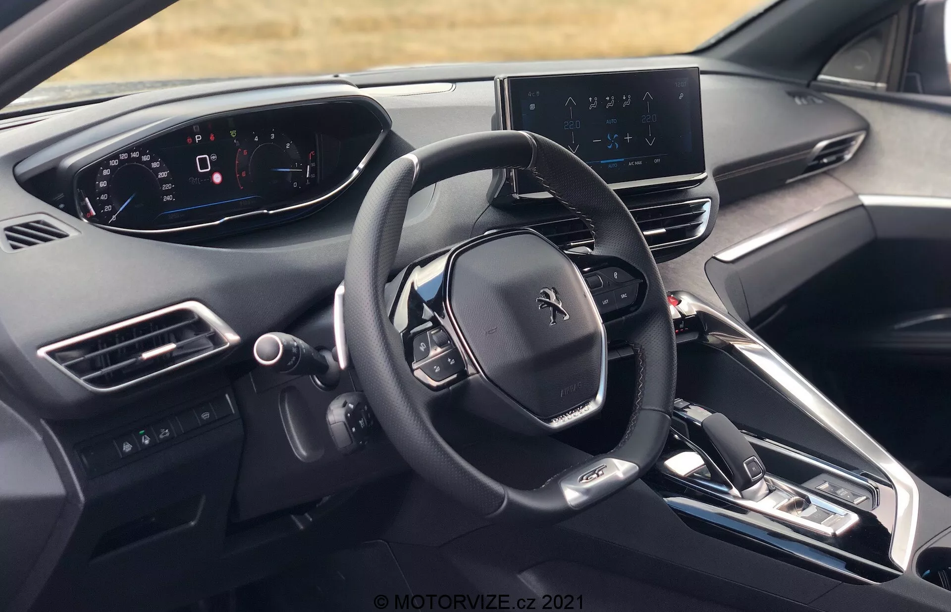 Widok wnętrza Peugeot 5008 2021 z perspektywy kierowcy, ukazujący kierownicę z logo Peugeot, wiele przycisków sterujących, w pełni cyfrowy zestaw wskaźników, centralny wyświetlacz systemu informacyjno-rozrywkowego z mapą nawigacji, elegancką deskę rozdzielczą z minimalną liczbą przycisków fizycznych, konsolę środkową z przyciskami do sterowania klimatyzacją i ustawieniami pojazdu, elektroniczną dźwignię zmiany biegów, kratki wentylacyjne, przycisk start/stop silnika, materiały wysokiej jakości i częściowy widok zagłówka siedzenia kierowcy.