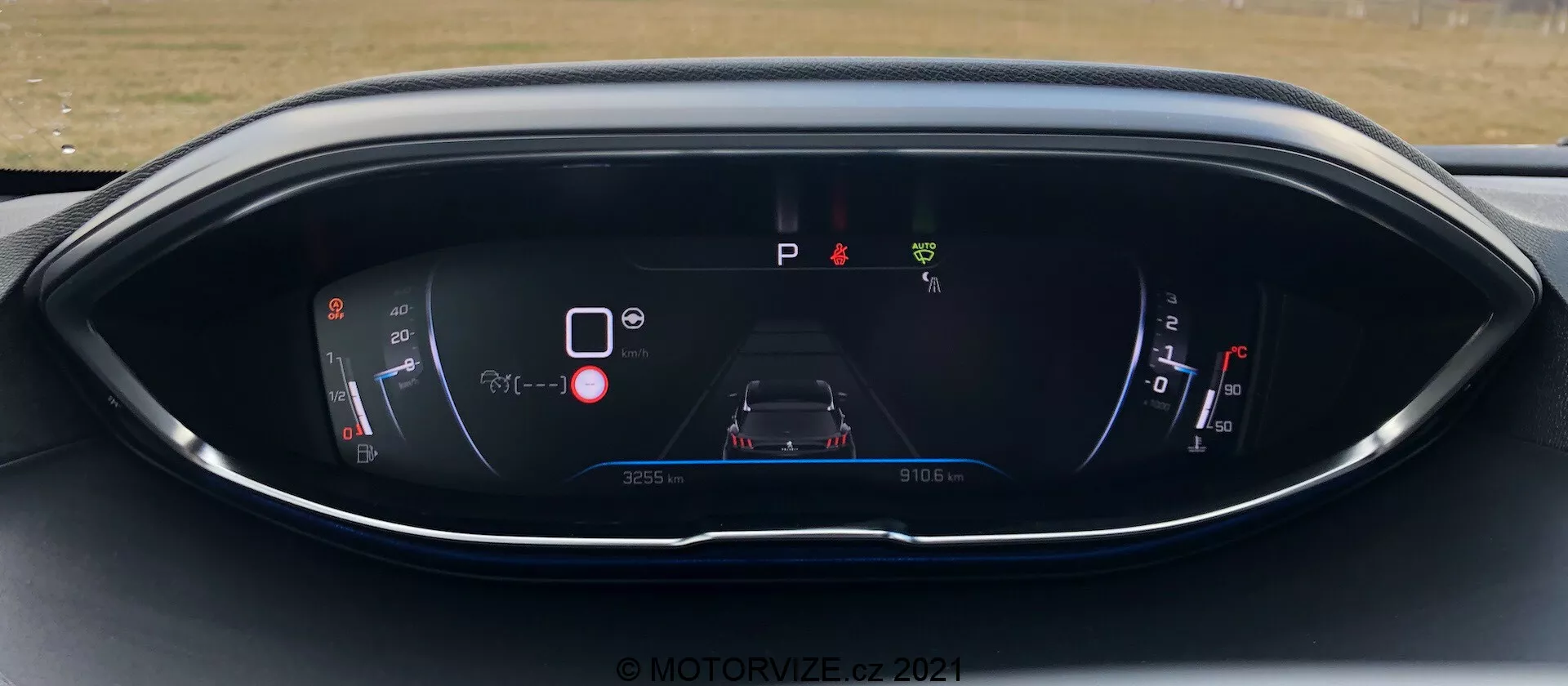 Zestaw wskaźników Peugeot 5008 2021, z cyfrowym wyświetlaczem z perspektywy siedzenia kierowcy. Wyświetlacz zawiera wskaźnik mocy/baterii, centralny grafik samochodu z ikonami ostrzegawczymi, wskaźnik 'P' trybu parkowania, wskaźnik paliwa i informacje o liczniku przebiegu/podróży. Wzornictwo obejmuje nowoczesny schemat kolorów niebiesko-czerwony na elegancko zakrzywionej obudowie. Znak wodny © MOTOR1ZED 2021.