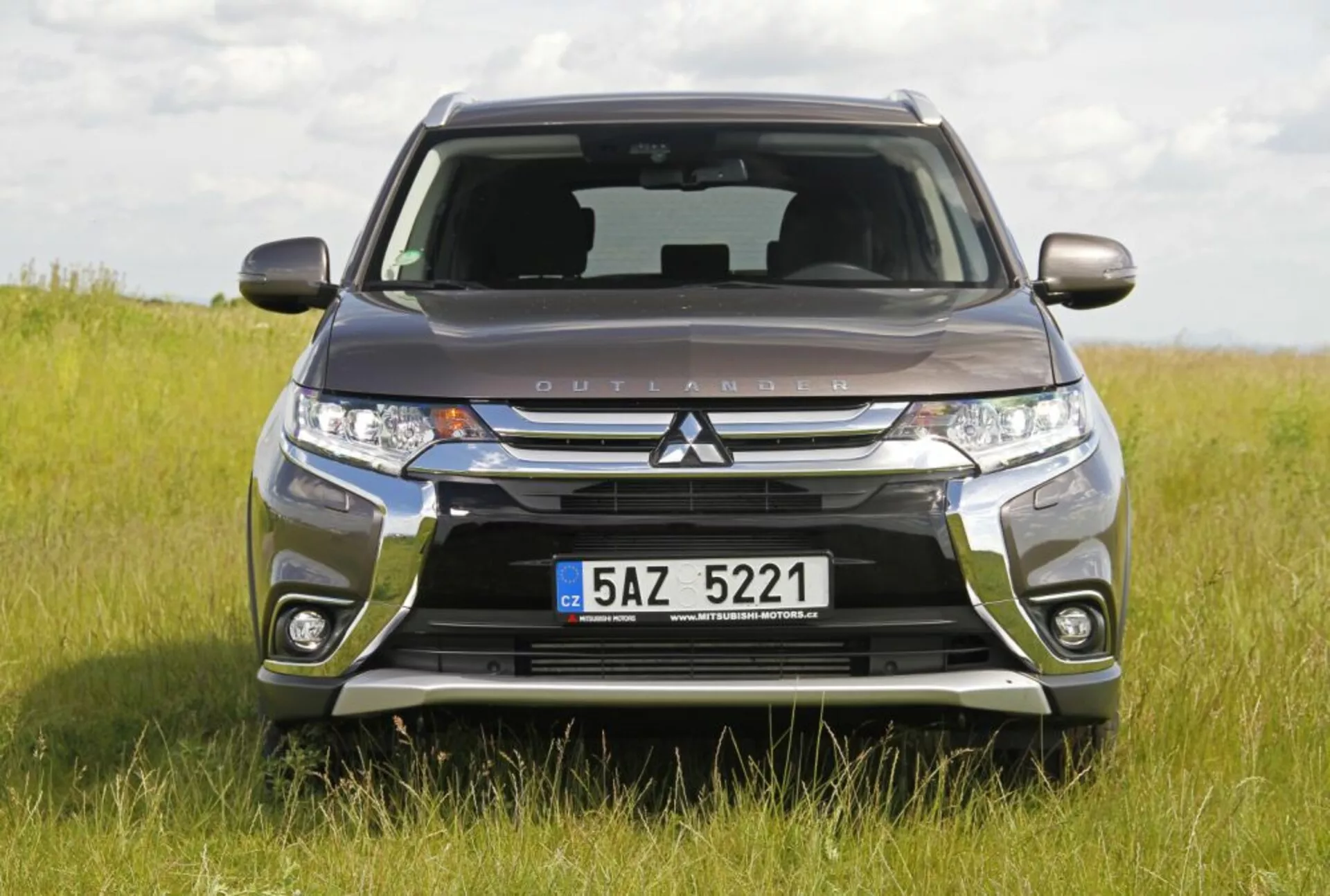 Zdjęcie czołowe Mitsubishi Outlander na trawiastym polu podkreślające jego grill Dynamic Shield, reflektory LED i solidną konstrukcję zderzaka, pokazujące zdolności terenowe i nowoczesne funkcje technologiczne w świetle dziennym.