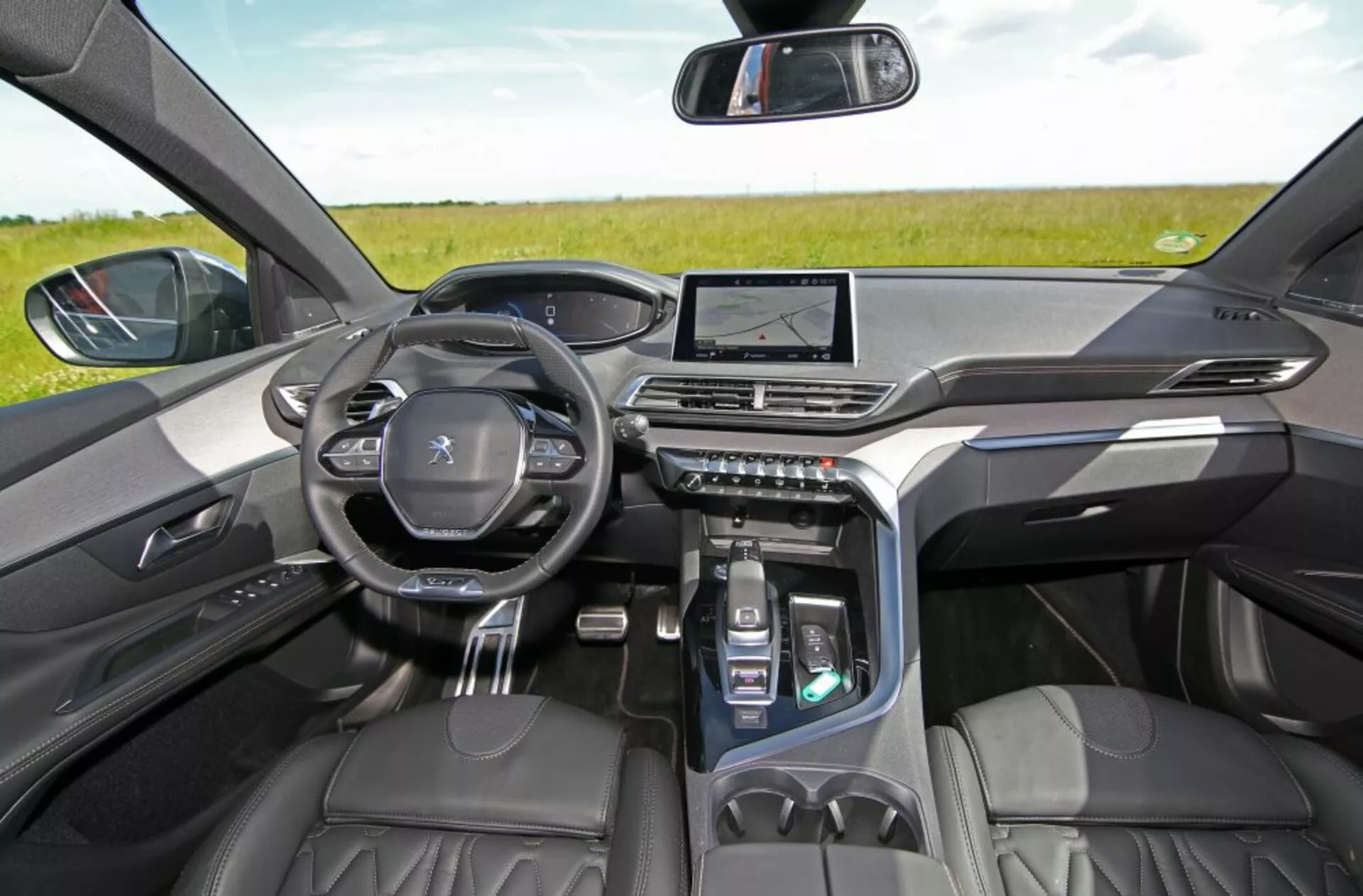 Widok wnętrza Peugeot 5008 z 2013 roku z tylnych siedzeń skupiający się na desce rozdzielczej, z przednimi siedzeniami z wysokiej jakości skóry, cyfrowym panelem wskaźników, kierownicą Peugeot z zamontowanymi sterowaniami, centralnym systemem infotainment z ekranem dotykowym i nowoczesnym centralnym panelem sterowania, w naturalnym świetle otoczenia