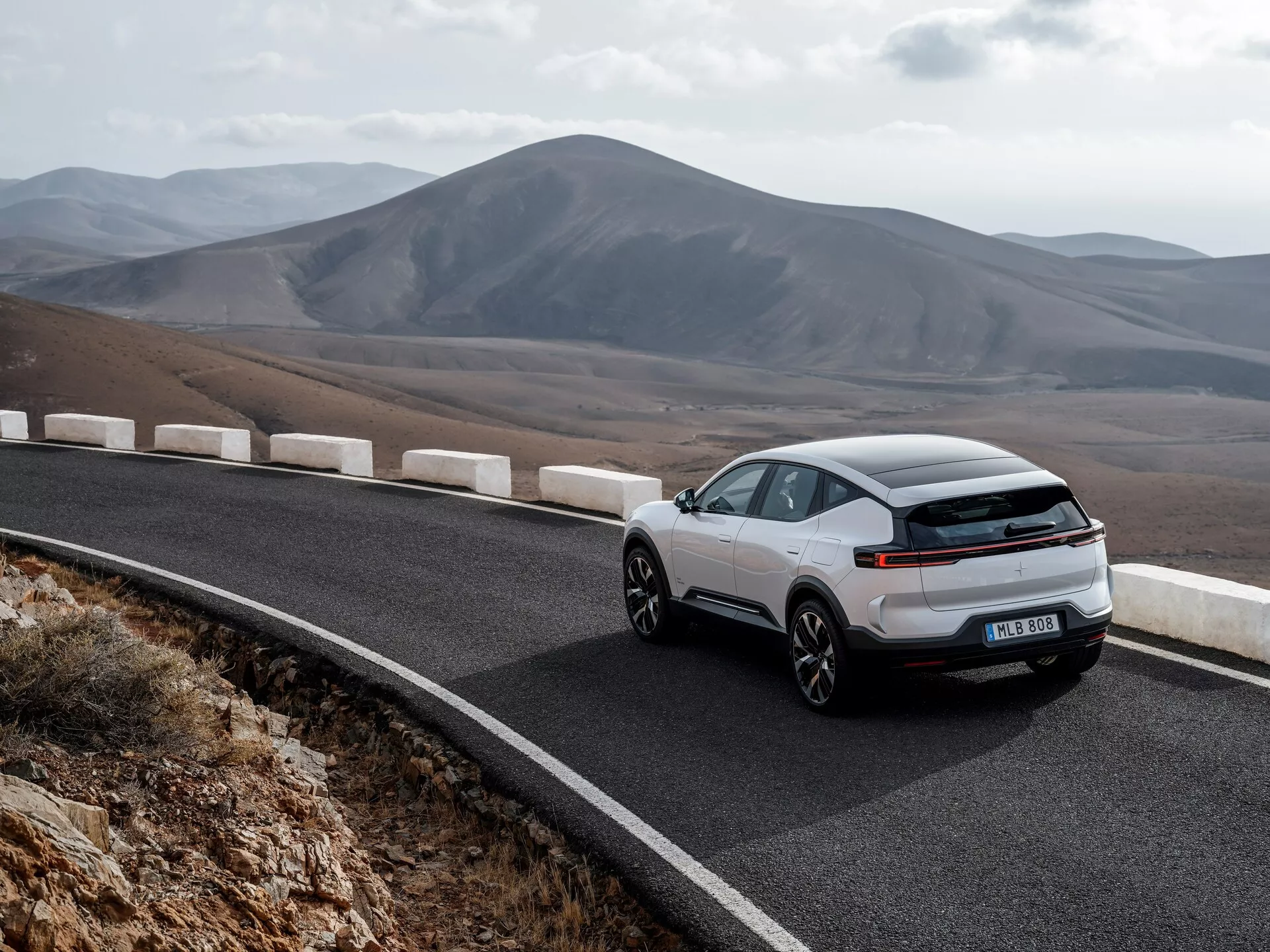 Polestar 3 1. generacji 2023 przedstawiony z dynamicznego trójkątnego widoku z tyłu na krętej górskiej drodze, podkreślający aerodynamiczną zewnętrzność, nowoczesny pasek świetlny LED i sportowy design typu coupe na tle malowniczych gór, podkreślający ekologiczną jazdę bez widocznej rury wydechowej, sugerując technologię elektryczną lub hybrydową.