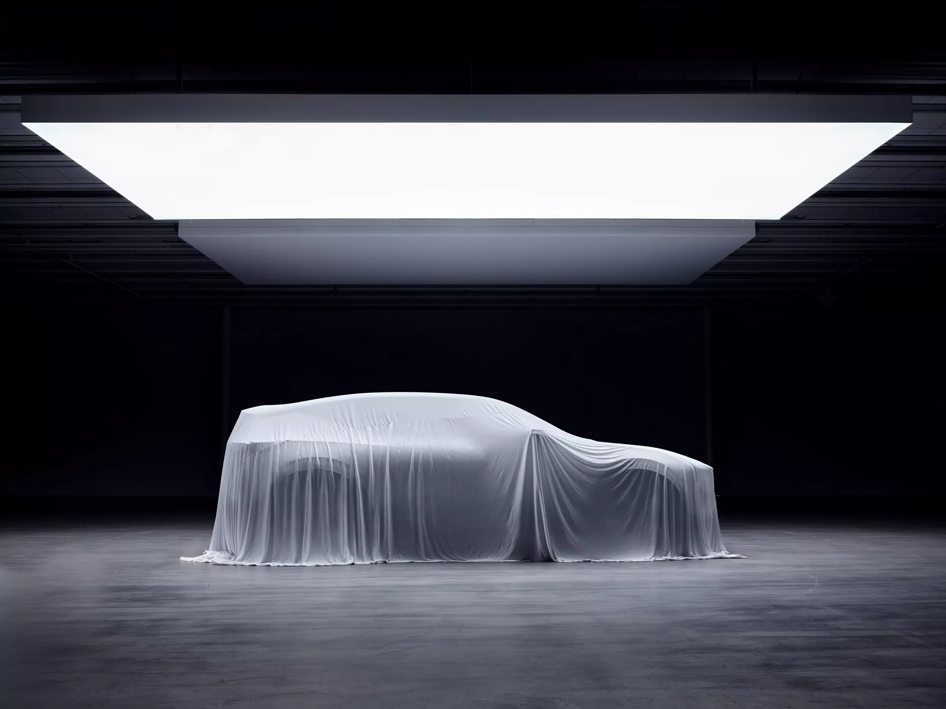 Odsłonięcie pierwszej generacji Polestar 3 2023 pod płótnem z profilu bocznego podkreślającego aerodynamiczny design i kontury coupe, pod dramatycznym oświetleniem salonu