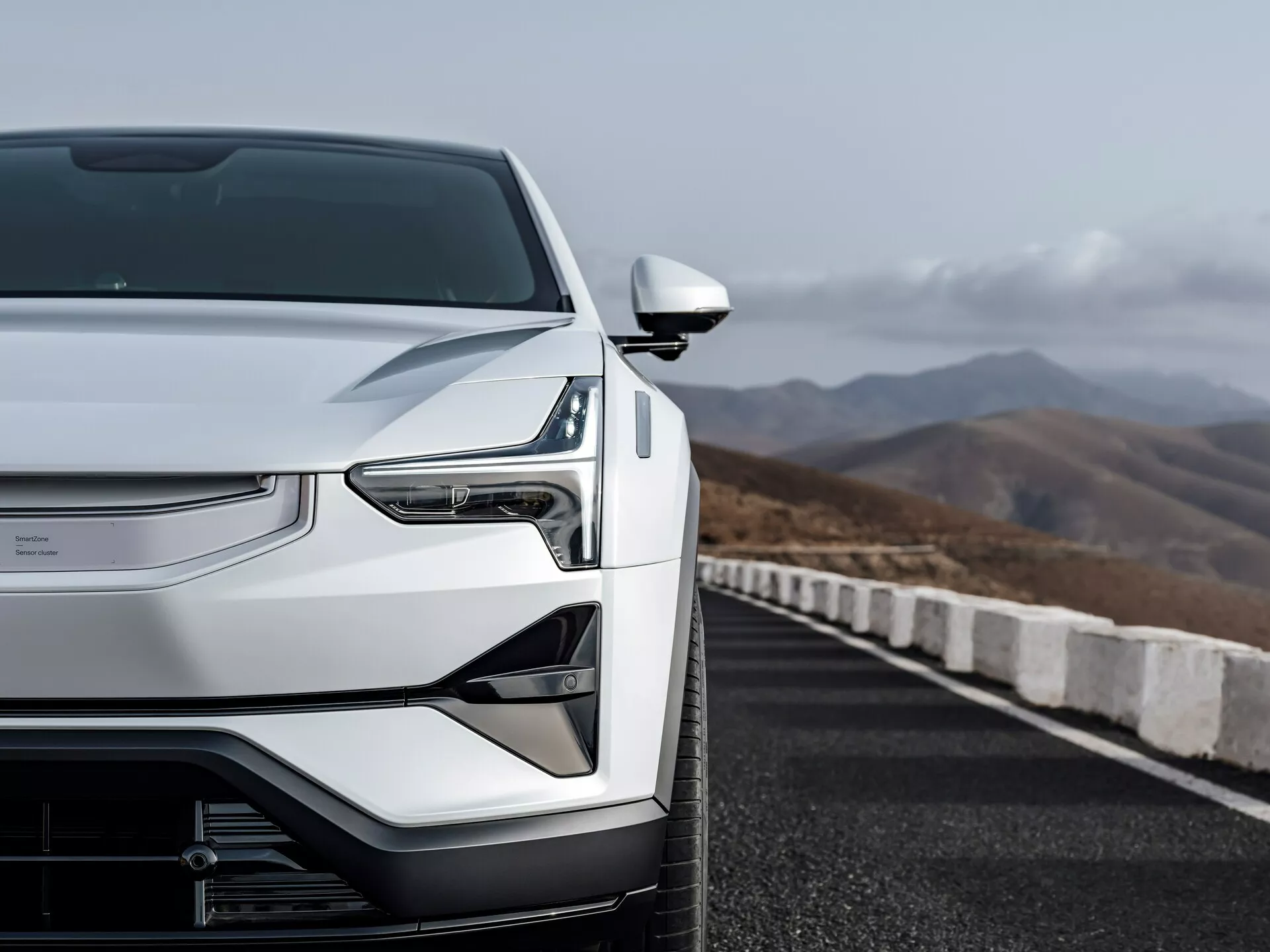 Widok z lewej przedniej strony Polestar 3 2023 pokazujący aerodynamiczny kaptur, zamkniętą kratę technologii pojazdu elektrycznego, reflektory LED i eleganckie lusterko boczne na drodze z górskim tłem