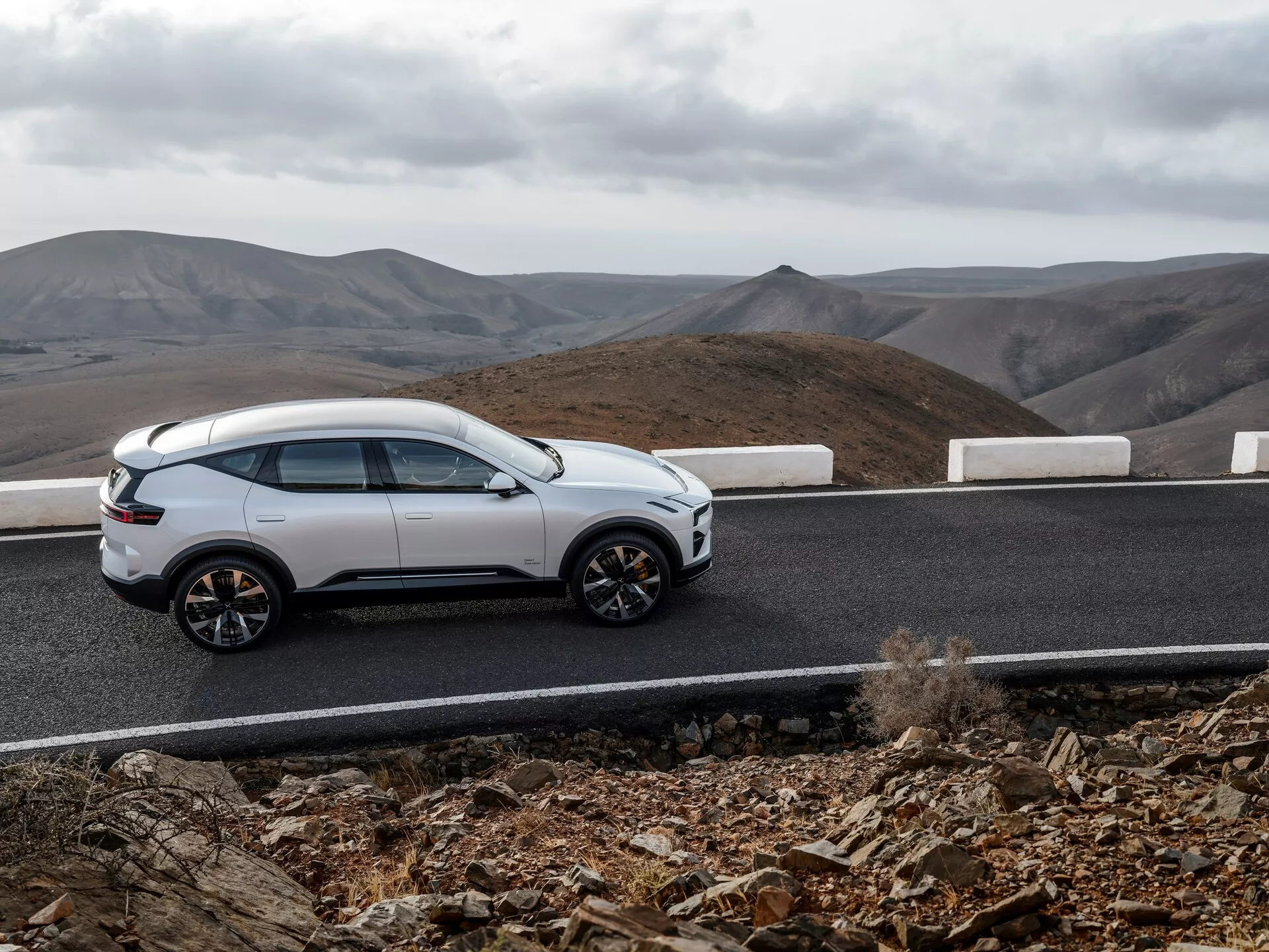 Biały SUV Polestar 3 z 2023 roku na zakrzywionej asfaltowej drodze z białym obramowaniem przeciwko zachmurzonemu, pagórkowatemu krajobrazowi, pokazujący profil boczny, elegancki design, koła z wieloramieniem i nowoczesne cechy jak chowane klamki i panoramiczny dach, sugerujący luksus i zdolności terenowe