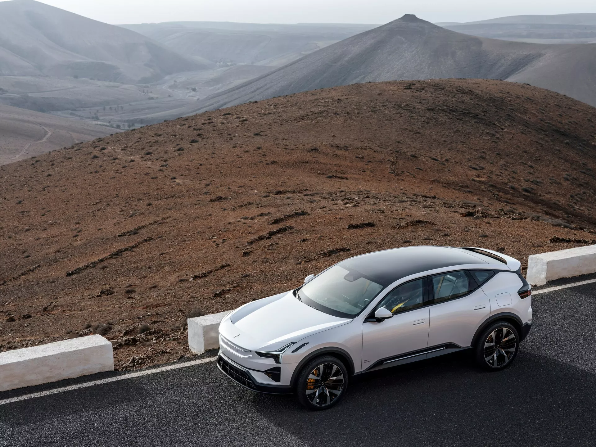 Nowoczesne SUV Polestar 3 1. generacji z 2023 roku zaparkowane przy drodze z jałowym krajobrazem wzgórz w tle, prezentuje aerodynamiczny design, panoramiczny dach, srebrne relingi dachowe, reflektory LED, sportowe koła o wielu szprychach, zaawansowaną technologię bezpieczeństwa, w spokojnym, odległym otoczeniu