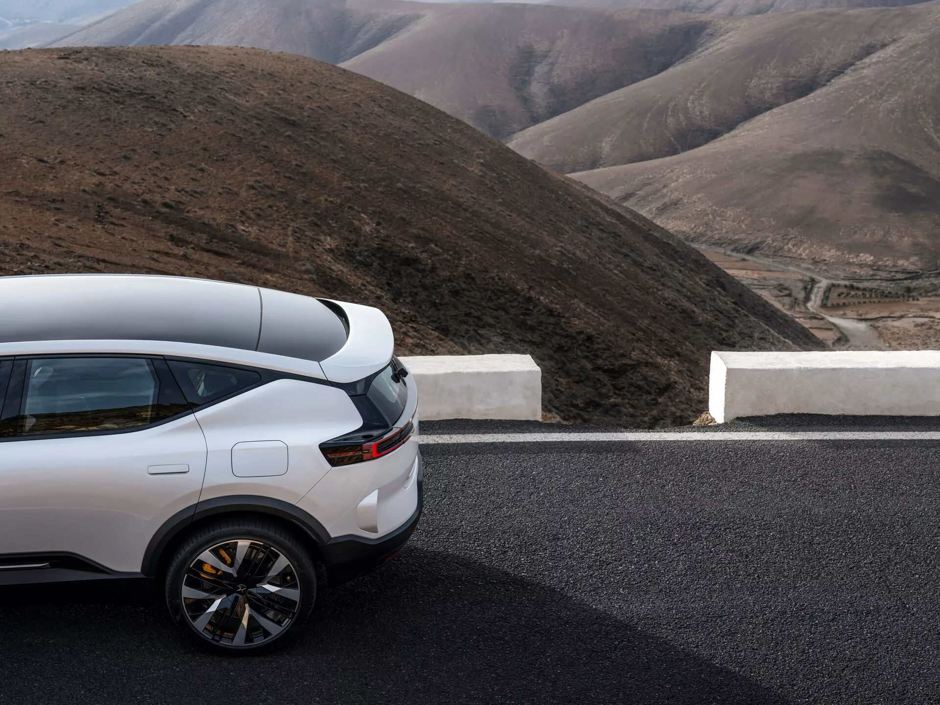 Polestar 3 pierwszej generacji z 2023 roku, prezentujący częściowy boczny i tylny widok na drodze, z wysokiej klasy białym SUV-em na tle górskim, podkreślającym sportowy tył, nowoczesny design lamp tylnych i luksusowy profil boczny