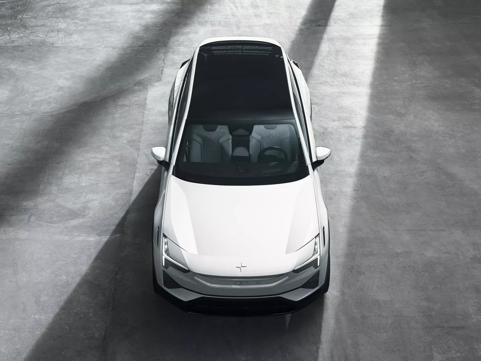 Sedan Polestar 3 pierwszej generacji z 2023 roku pokazany z wysokiego kąta frontowego w środowisku salonu, podkreślający jego nowoczesny design, biały kolor i cechy przednie, w tym maskę, dach, przednią szybę, drzwi przednie, eleganckie reflektory LED i grill z niewidocznym znaczkiem, sugerujący możliwe cechy pojazdu elektrycznego