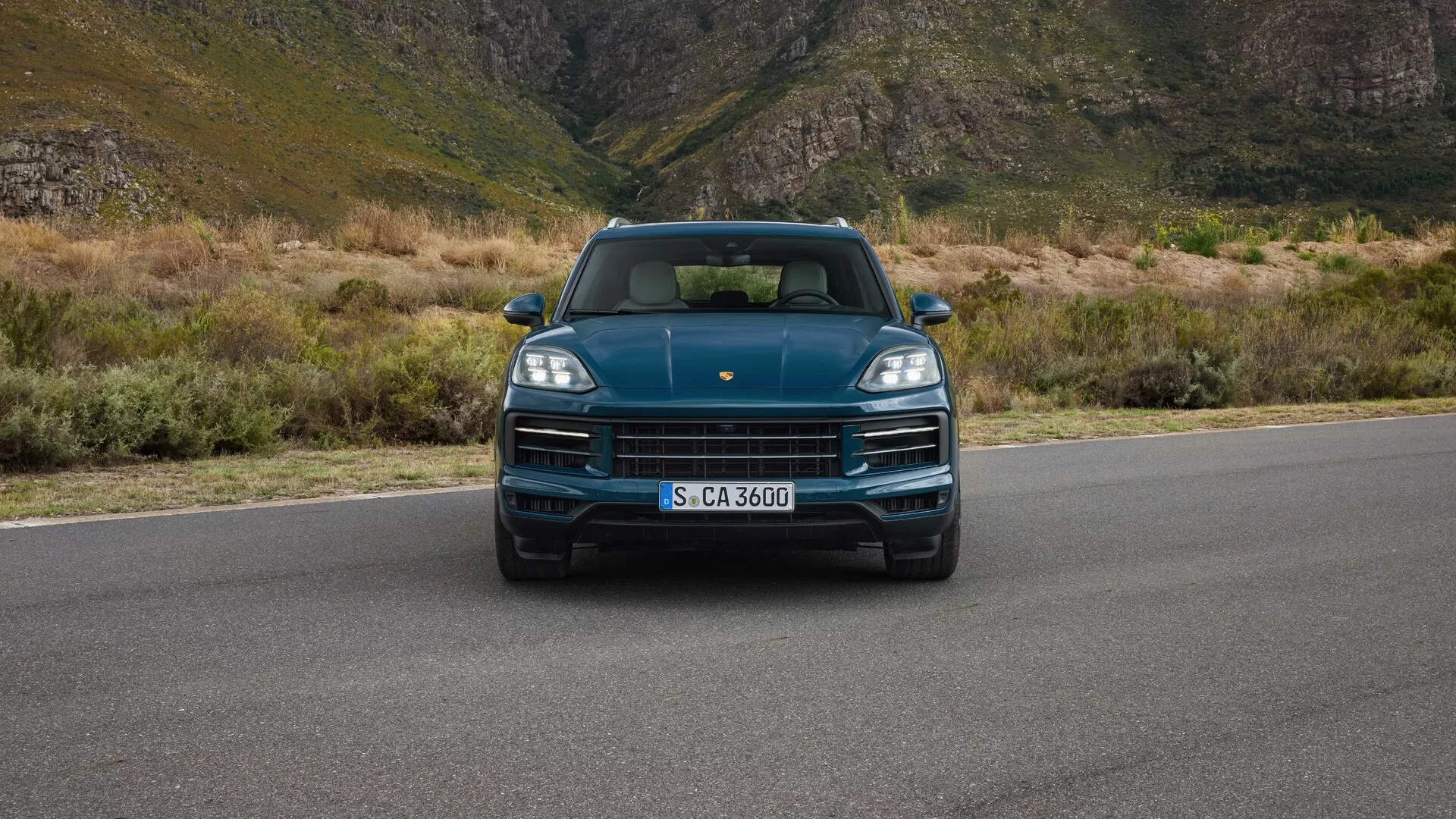Porsche Cayenne 2023 (9YA Facelift) przedstawiony z przodu, na wysokości oczu, prezentujący reflektory LED, wyeksponowany grill z logotypem marki, sportowy design maski i zintegrowane czujniki parkowania, na tle górskiego krajobrazu, co sugeruje luksus i przygodę