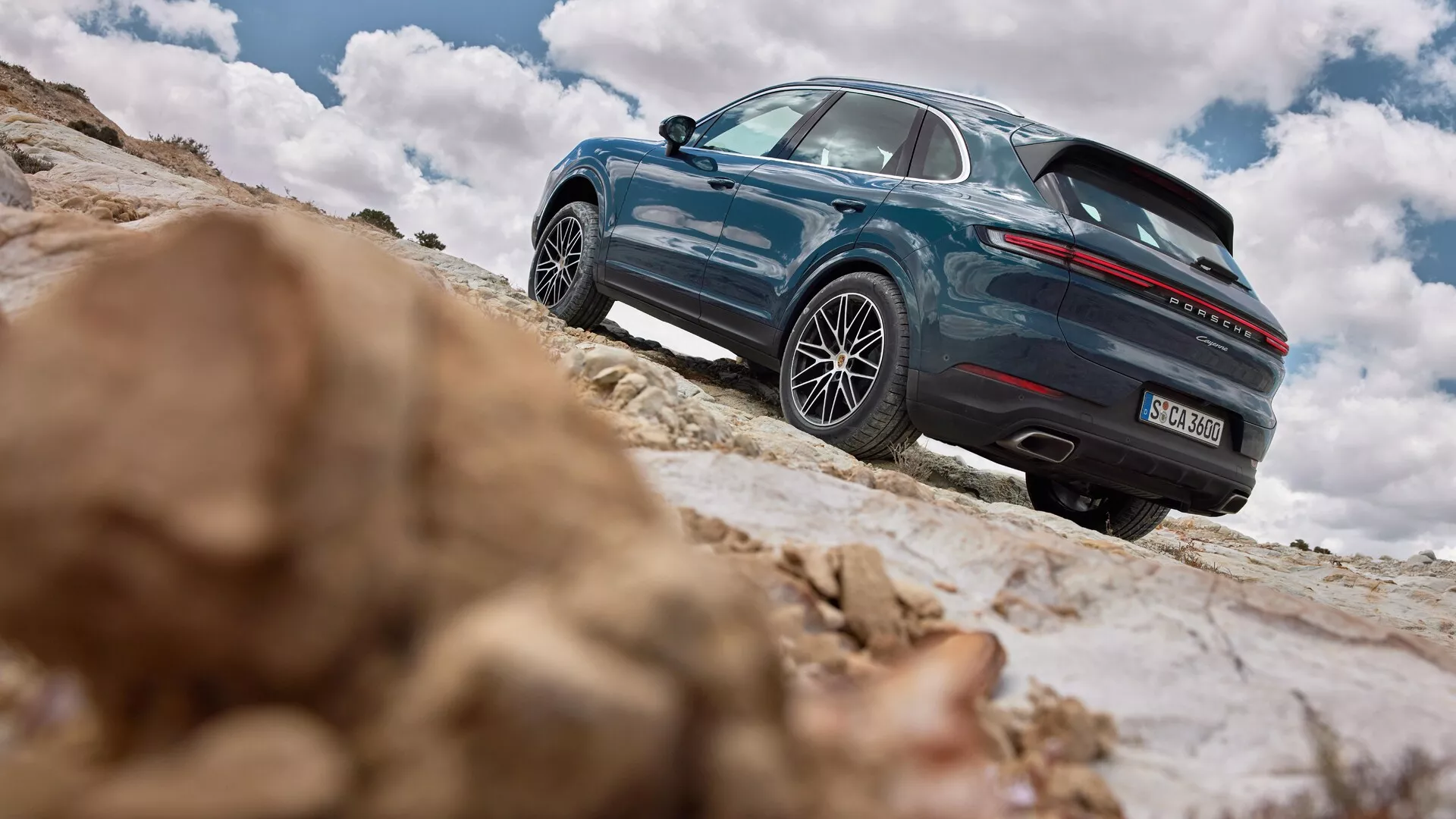 Porsche Cayenne 2023 (9YA-facelift) zaprezentowane na nachylonym skalistym terenie podkreślające jego zdolności terenowe, pokazujące lewy tylny profil z detalami designu kół, podwójnych wydechów i spoileru tylnego na tle dramatycznego nieba