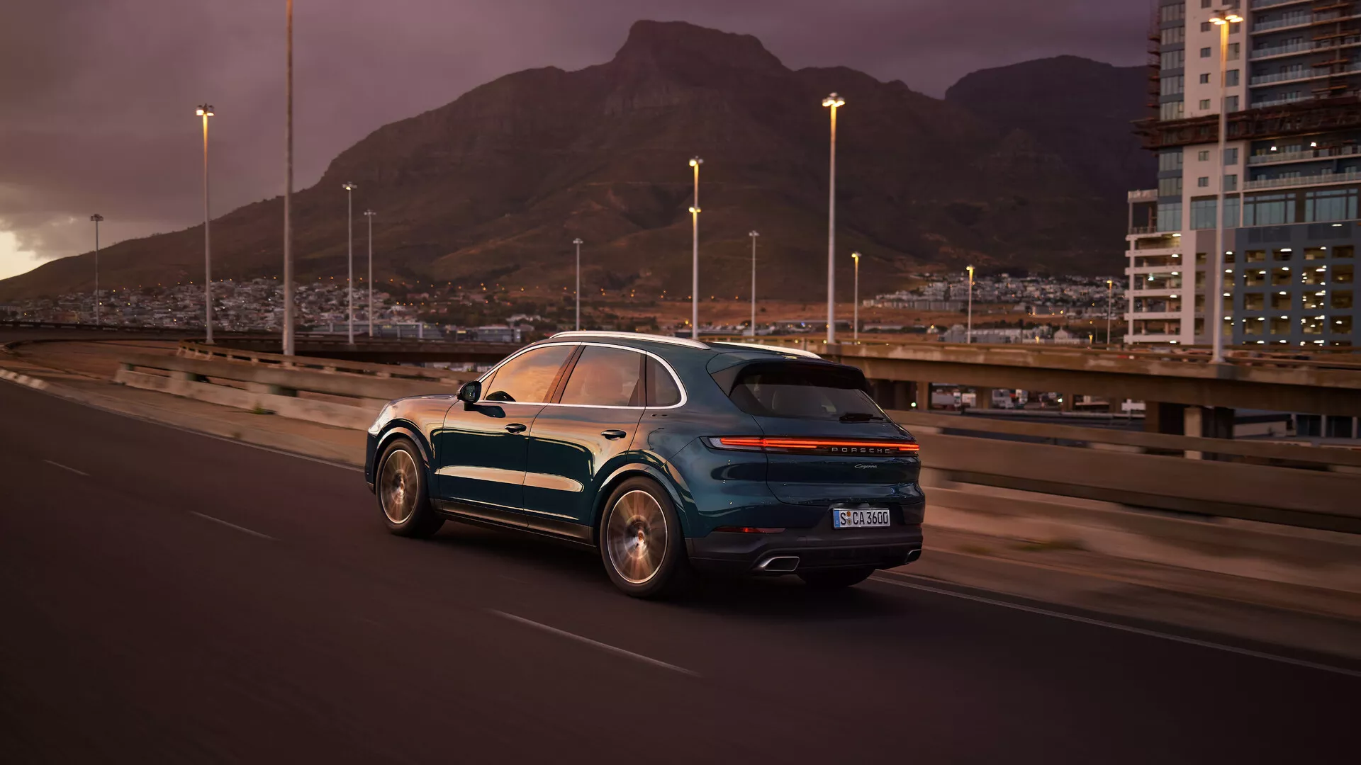 SUV Porsche Cayenne 2023 (9YA-facelift) jedzie po autostradzie o zmierzchu, prezentując widok z tyłu trzy czwarte z metalicznym lakierem, felgami aluminiowymi, LEDowymi światłami tylnymi, podwójnym wydechem i panoramicznym dachem na tle miejskiego krajobrazu z malowniczym pasmem górskim.