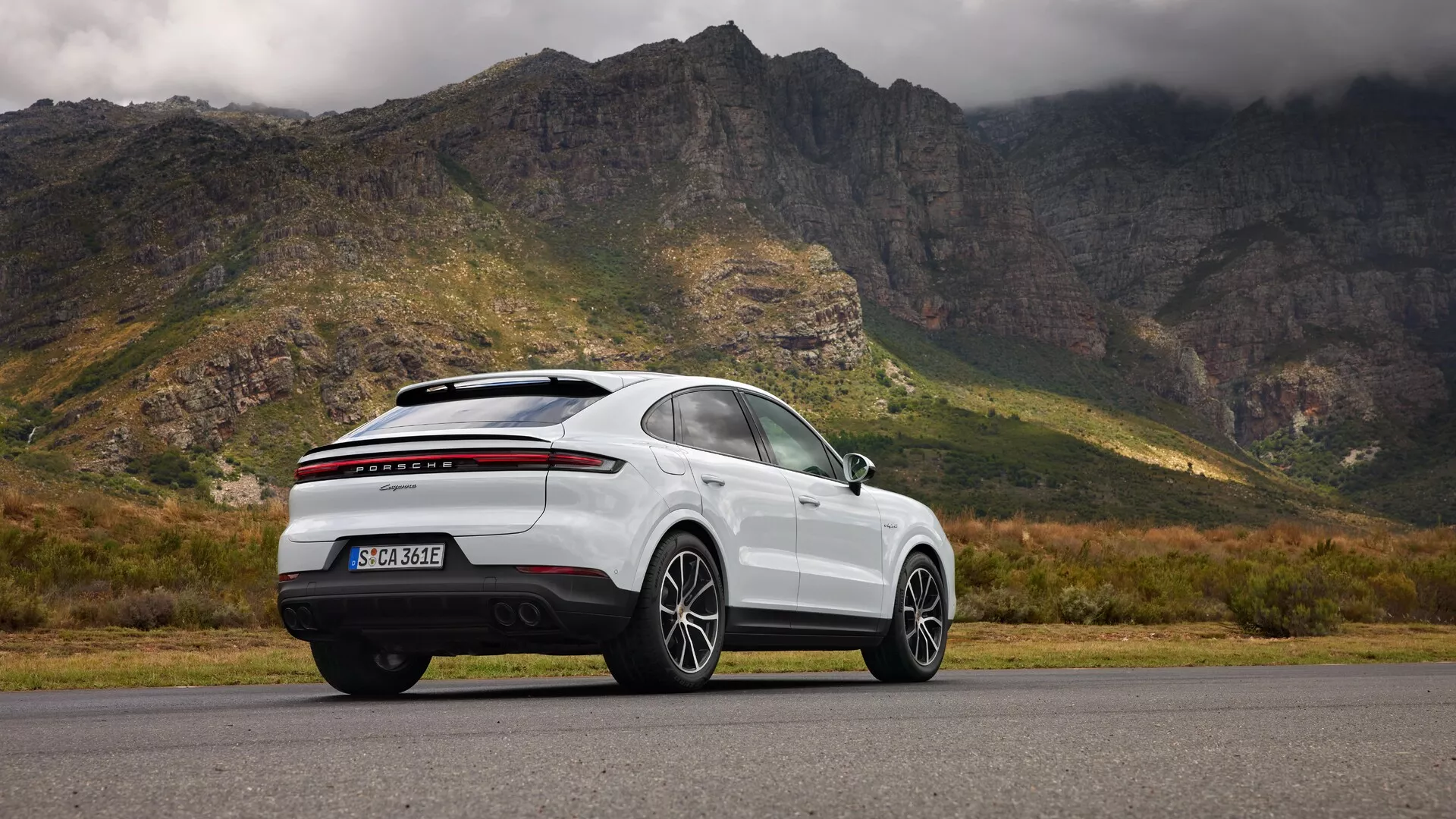 Nowoczesne białe SUV typu crossover Porsche Cayenne (9YA Facelift) z 2023 roku fotografowane z boku-tyłu na tle dramatycznego krajobrazu górskiego z zachmurzonym niebem, ukazujące aerodynamiczny profil pojazdu, wieloramienne felgi aluminiowe, elegancką linie dachu ze zintegrowanym tylnym spojlerem, panoramiczny dach, diody LED na tylnych światłach, i sugerowane zaawansowane funkcje technologiczne