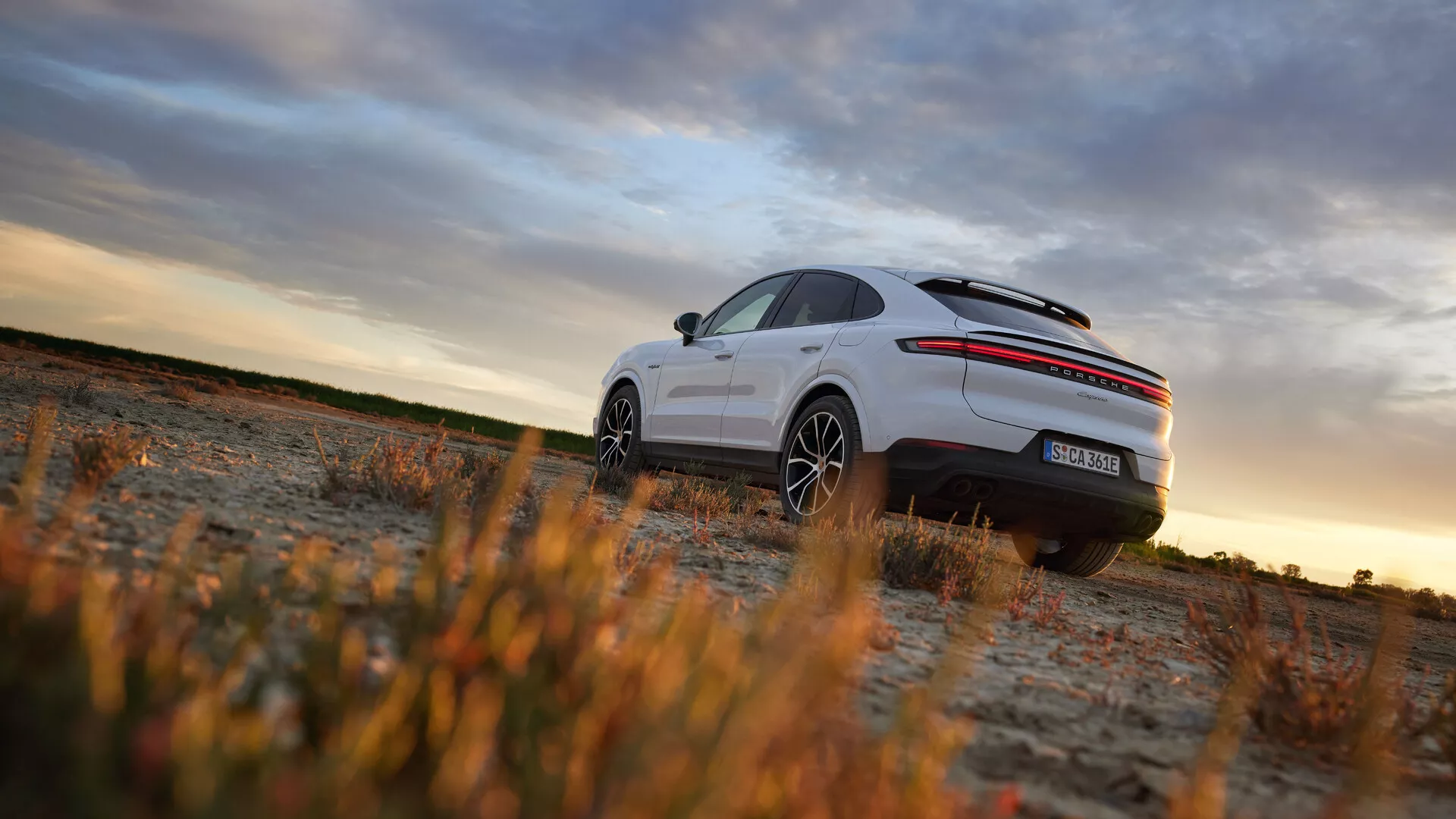Porsche Cayenne 2023 (lifting 9YA) uchwycone z niskiego kąta w wiejskim otoczeniu podczas złotej godziny, przedstawiające sportową postawę, tylne lampy LED, podwójny wydech, spoiler dachowy, duże felgi aluminiowe i możliwy pas projektu specjalnej edycji.