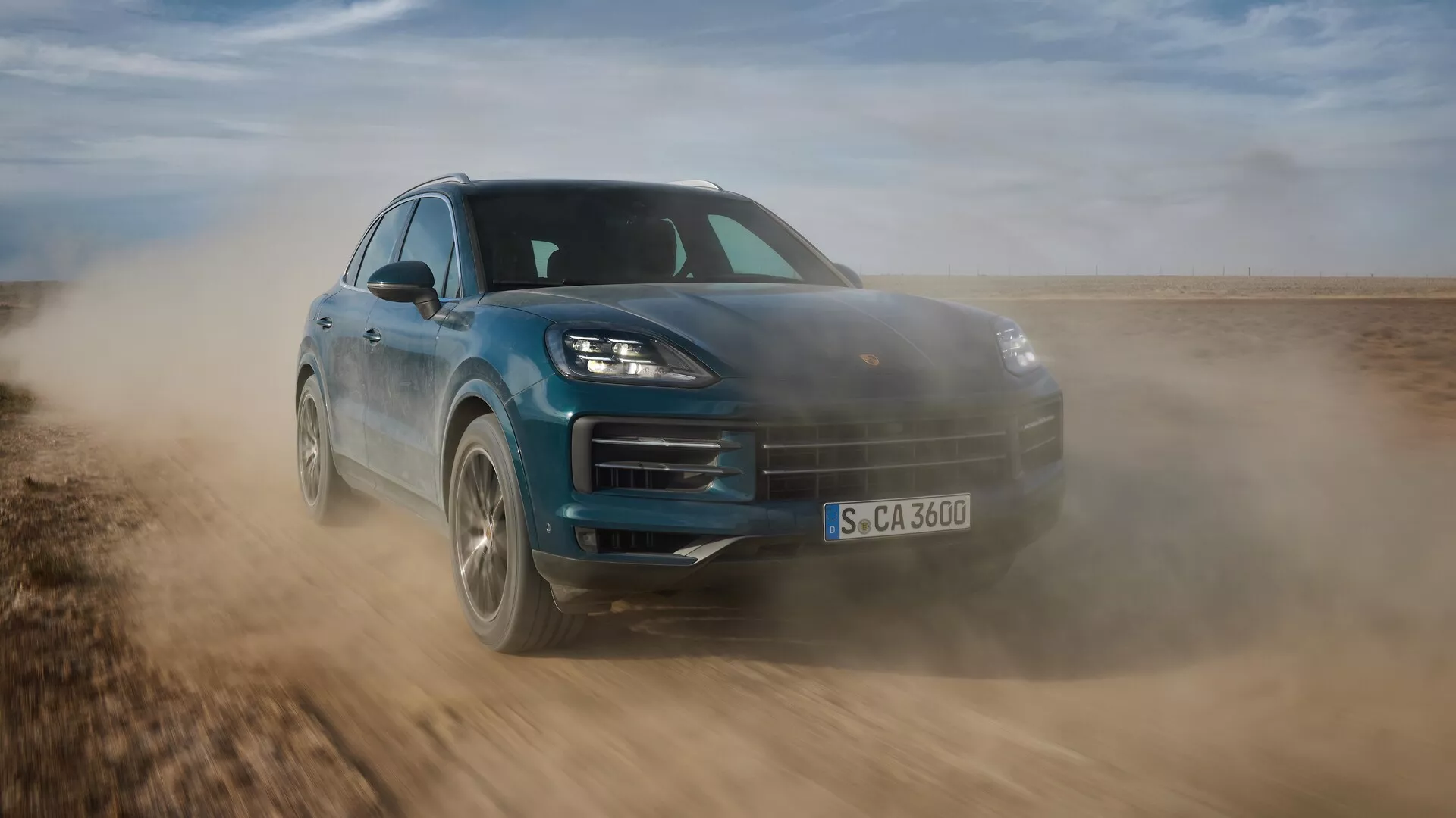 Porsche Cayenne 2023 (lifting 9YA) w dynamicznym ruchu, prezentujące swój elegancki design i zdolności terenowe, uchwycone z niskiego, przedniego trzy-czwartego kąta podkreślającego sportową postawę, zaawansowane reflektory LED i cechy aerodynamiczne na nieutwardzonej nawierzchni.