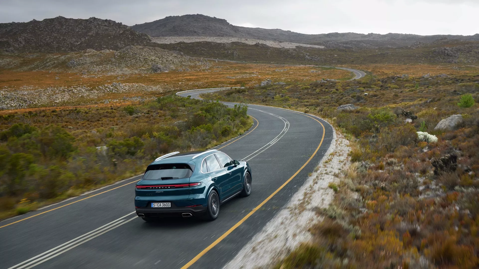 Porsche Cayenne 9YA-Facelift 2023 jadący krętą drogą przez górzysty teren z dynamicznym widokiem z tyłu z trzech czwartych podkreślającym sportowy design, charakterystyczne tylne światła, tylny spoiler, podwójne porty wydechowe i zaawansowaną technologię na tle surowego krajobrazu pod pochmurnym niebem