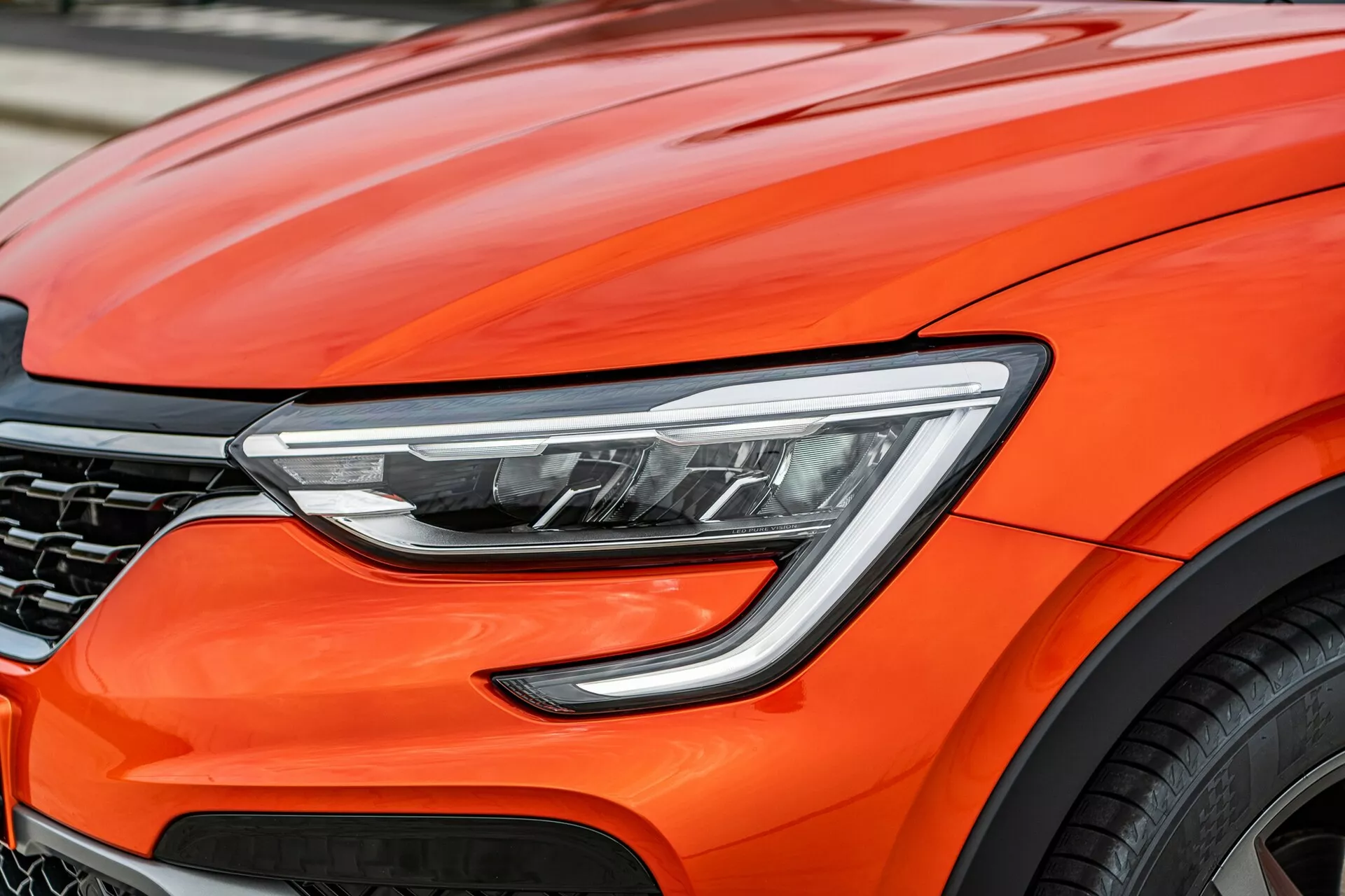 Zbliżenie przedniej części jaskrawo pomarańczowego Renault Arkana 2021, skupiające się na nowoczesnych detalach projektowych, w tym zaawansowanych reflektorach LED, aerodynamicznych krzywiznach maski, częściowo widocznej ciemnej kratce w kształcie plastra miodu oraz fragmencie dużego aluminiowego koła, sugerujących wydajność premium i zaawansowaną technologię motoryzacyjną.