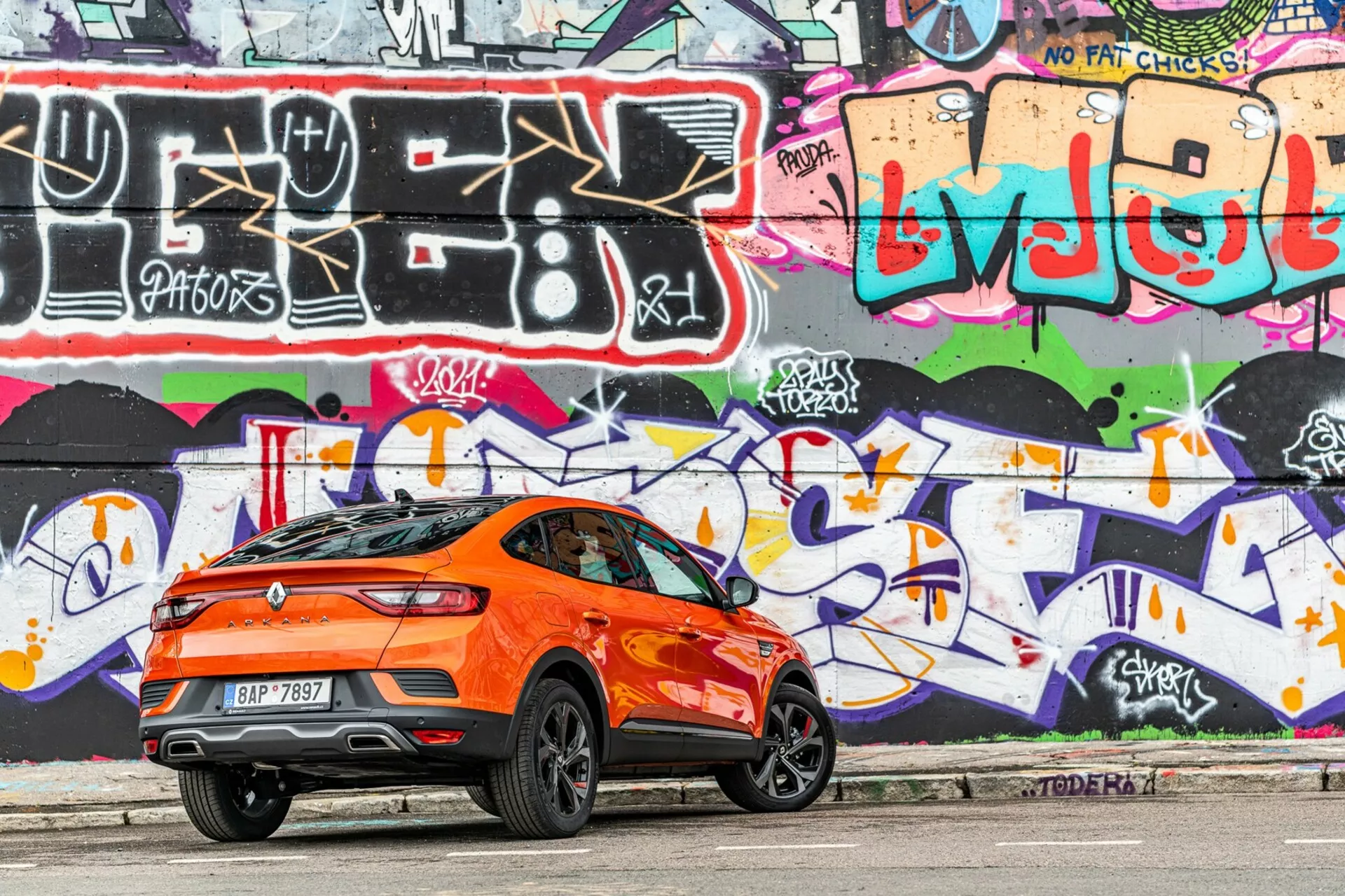 Widok profilu bocznego jasnopomarańczowego crossovera Renault Arkana 2021 zaparkowanego na miejskiej drodze. Zdjęcie wykonane od prawej strony z naciskiem na tylne trzy czwarte, uchwyca sportową, opadającą linię dachu, tylny spojler, wysoką linię nadwozia, duże stylowe felgi aluminiowe, przyciemniane szyby oraz aerodynamiczne linie charakterystyczne. Tylne światła są eleganckie i prawdopodobnie LED. Tło przedstawia intensywnie pomalowaną graffiti ścianę, co podkreśla miejski charakter. Zdjęcie wykonane w świetle dziennym, pokazuje delikatne cienie pod samochodem, podkreślając szczegóły designu.