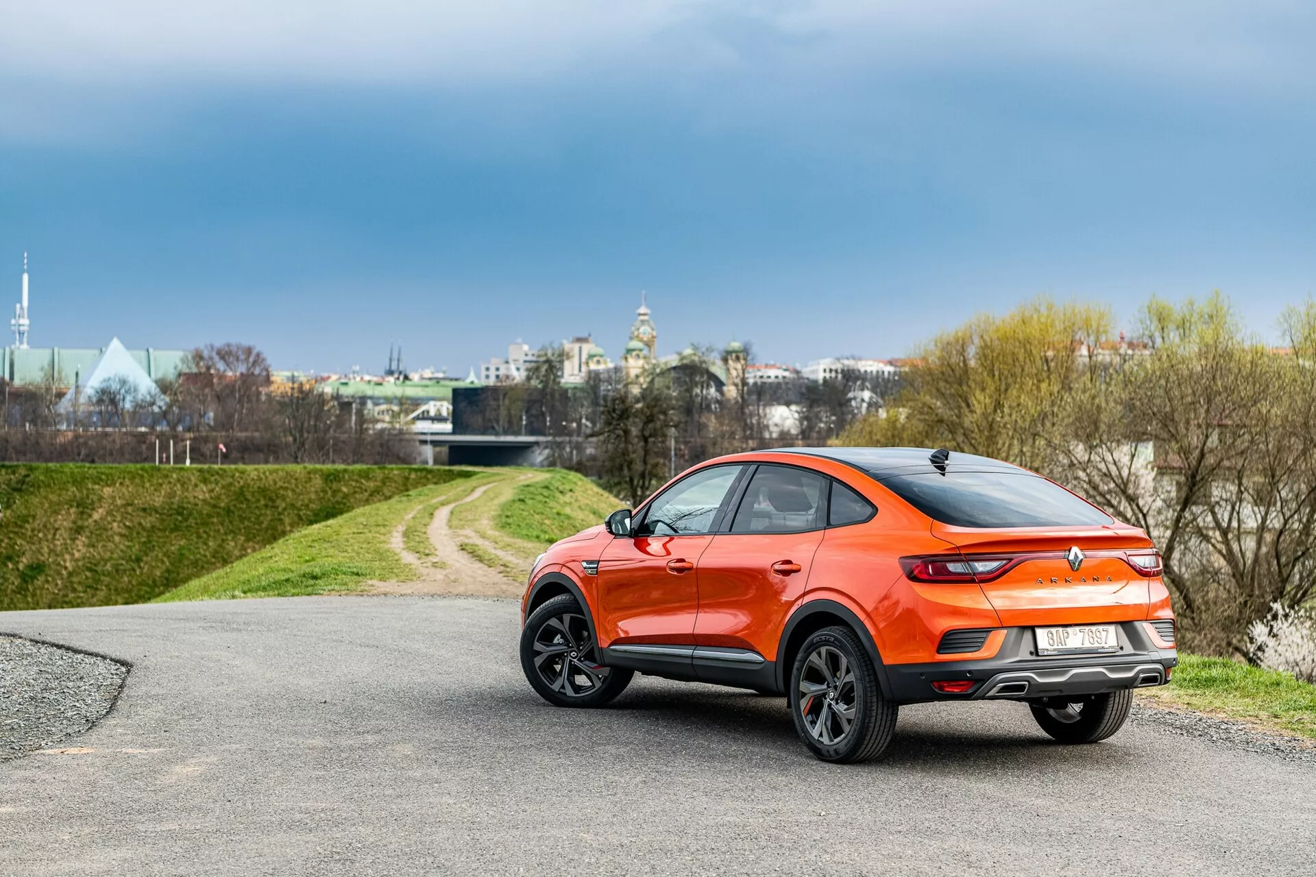 Widok trójczwartalny tylnego rogu hatchbacka Renault Arkana 2021 z sportowym designem crossover w żywym metalicznym pomarańczowym kolorze. Samochód posiada pochylony dach, czarne plastikowe osłony wokół nadkoli, spojler na wierzchu bagażnika, eleganckie tylne światła LED i duże wieloramienne felgi aluminiowe. Jest umieszczony na asfaltowej drodze prowadzącej do trawiastego terenu, pod zachmurzonym niebem w pobliżu miejskiego, ale spokojnego otoczenia.