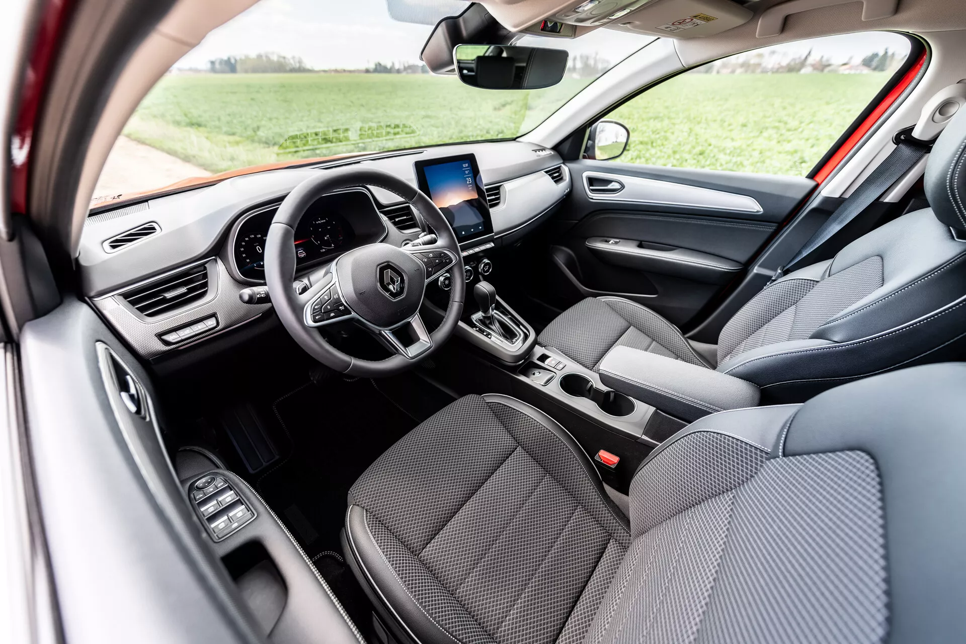 Wnętrze Renault Arkana 2021 prezentujące przednie siedzenia z szarej tkaniny, wielofunkcyjną kierownicę z trzema ramionami z wbudowanymi przyciskami sterującymi, cyfrowy wyświetlacz deski rozdzielczej, ekran systemu infotainment z nawigacją GPS nad centralnymi nawiewami, manualne sterowanie klimatyzacją, automatyczną dźwignię zmiany biegów i kontrolki elektrycznych szyb na drzwiach kierowcy, oświetlone naturalnym światłem dziennym w otoczeniu zewnętrznym