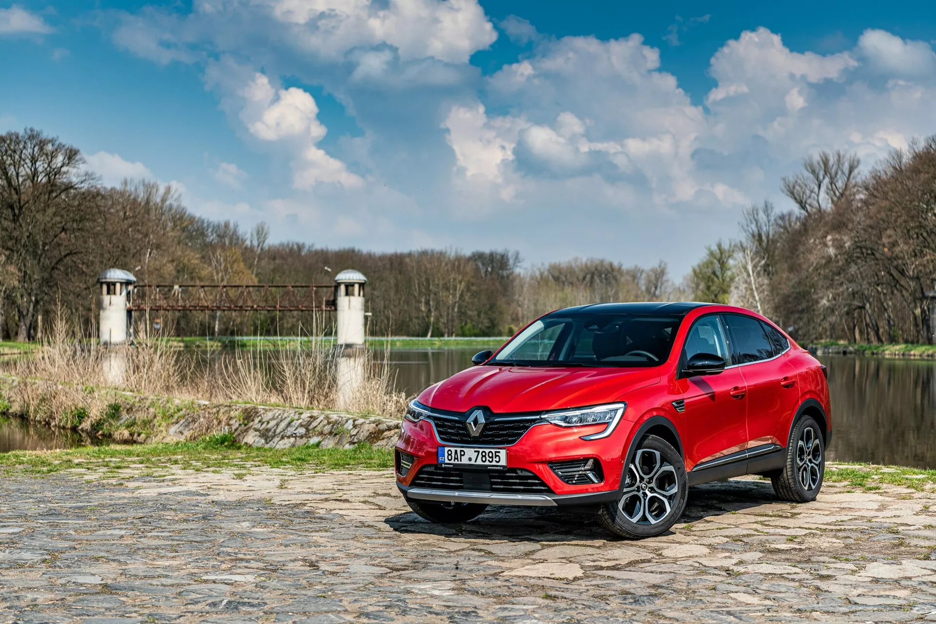 Na zdjęciu widoczny jest Renault Arkana 2021, zaparkowany na brukowanej ścieżce w pobliżu rzeki lub jeziora, z roślinnością i strukturami przypominającymi wieże tamy lub mostu w tle. Samochód jest uchwycony z niskiego kąta, w ujęciu trzech czwartych z przodu, podkreślając przednią kratkę, reflektory LED oraz część profilu bocznego, w tym drzwi kierowcy i przedni błotnik. Nowoczesne cechy designu, takie jak wyrazista sygnatura świetlna LED i duże aluminiowe koła, sugerują wyższy poziom wyposażenia. Odbijająca światło metaliczna farba dodaje walorów estetycznych, podczas gdy ukryte sensory wspomagania kierowcy oraz technologia wbudowana w zderzaki i boczne lusterka zwiększają funkcjonalność pojazdu.