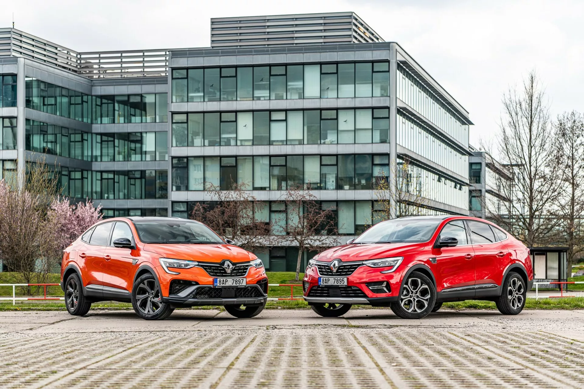 Obraz przedstawia dwa pojazdy w stylu SUV zaparkowane obok siebie przed nowoczesnym budynkiem biurowym ze szklaną fasadą. Pojazd po lewej stronie jest pomalowany na jaskrawy pomarańczowy kolor z metalicznym połyskiem i ma charakterystyczną przednią kratkę, ostre reflektory LED, rzeźbioną maskę oraz duże wieloramienne felgi aluminiowe z niskoprofilowymi oponami. Samochód po prawej stronie ma metaliczne czerwone wykończenie, z podobną ogólną sylwetką, ostrymi reflektorami, wyróżniającą się kratką i stylowymi dużymi felgami aluminiowymi. Oba pojazdy wyróżniają się kanciastymi liniami, aerodynamicznymi lusterkami i kontrastującymi dolnymi wykończeniami. Zdjęcie, wykonane pod niskim kątem blisko poziomu gruntu, podkreśla miejskie otoczenie i podkreśla projekt, obecność i zaawansowane funkcje technologiczne pojazdów.