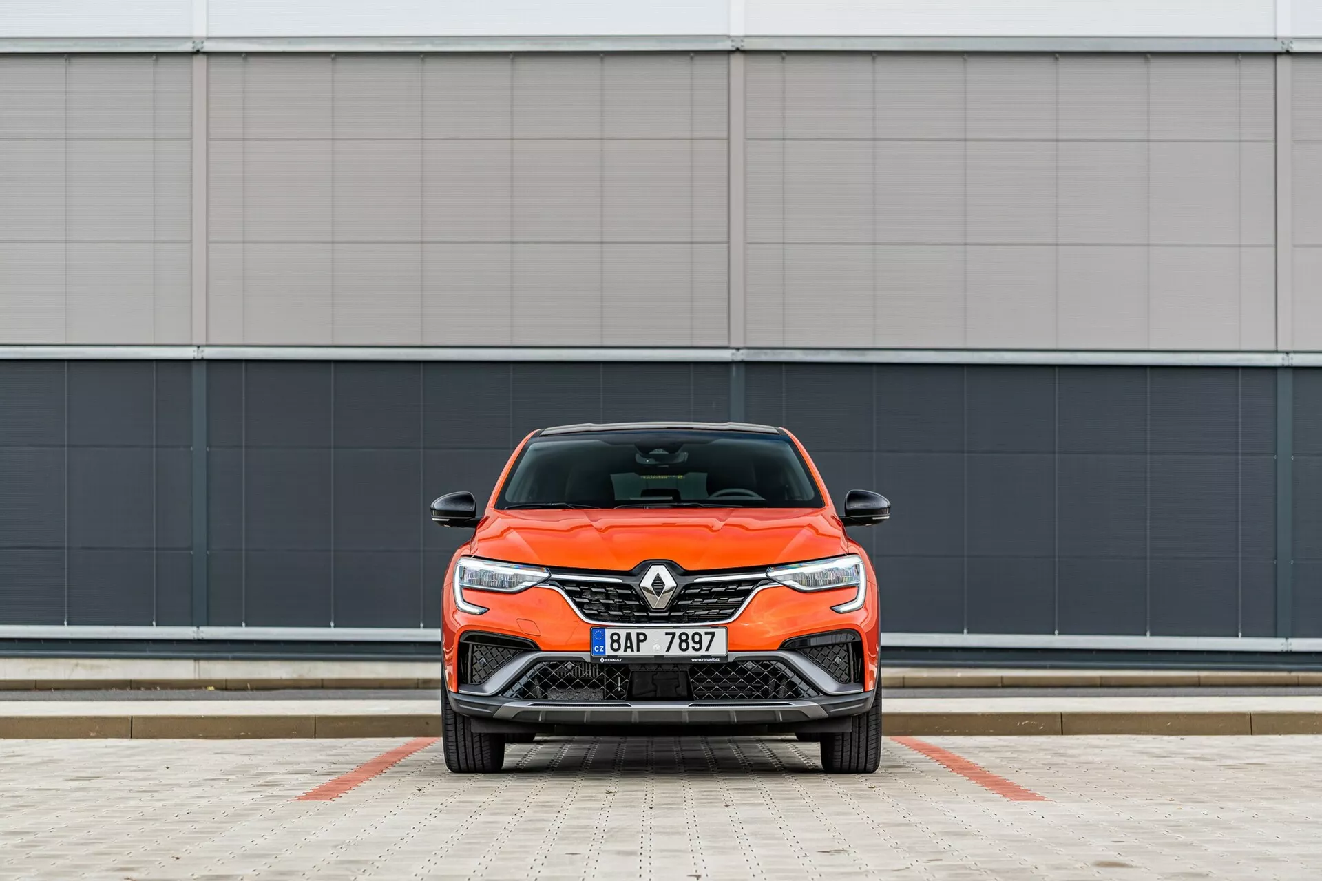Widok z przodu jasno pomarańczowego samochodu marki Renault, podkreślającego jego nowoczesny design ze smukłymi reflektorami i przednim grillem, zaparkowanego prostopadle do kamery na tle minimalistycznej szarej ściany w wybrukowanym miejscu parkingowym.