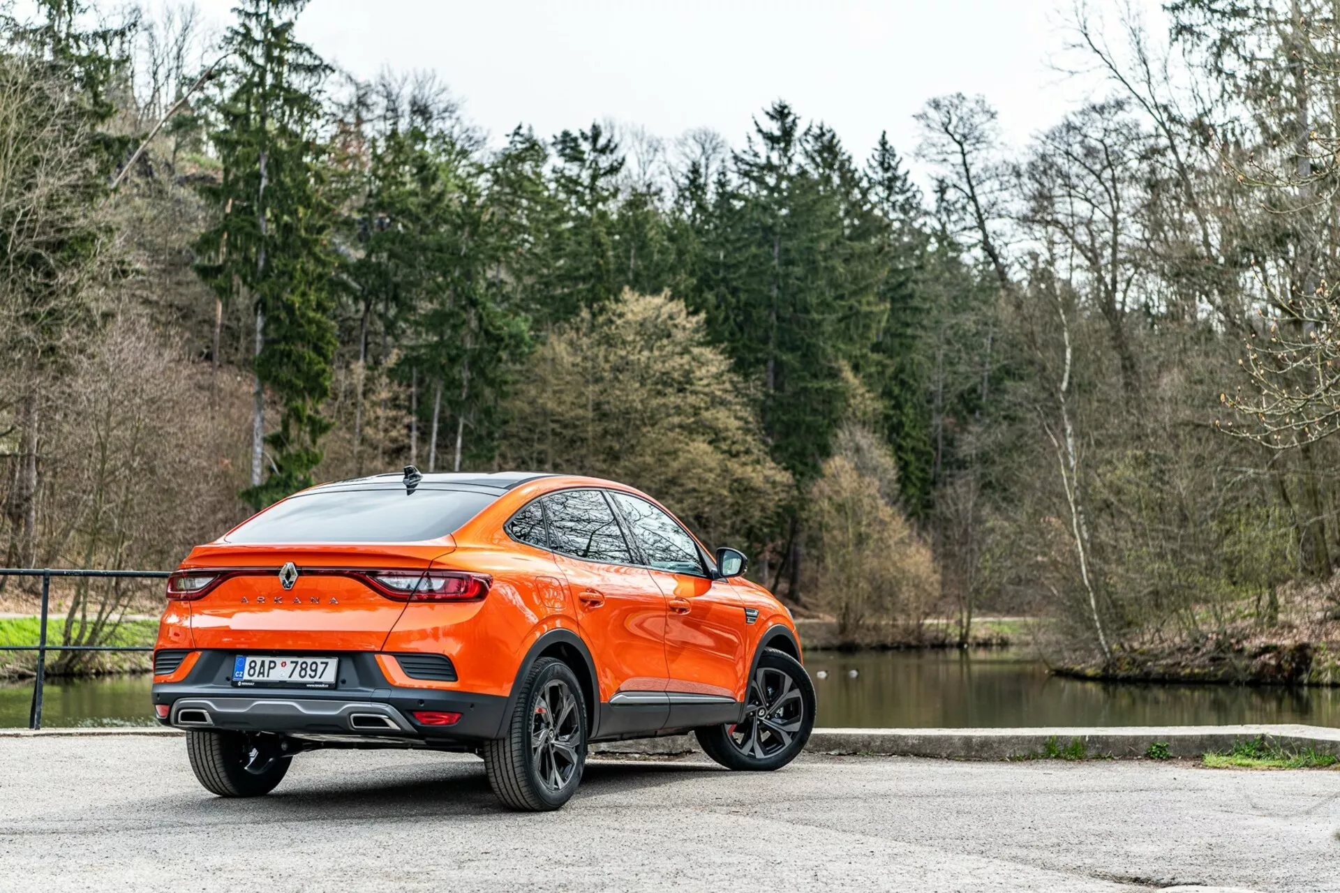 Renault Arkana 2021 widziany z tyłu z kąta trzech czwartych, szczegółowy widok bocznego i tylnego profilu, prezentujący nowoczesny crossover/SUV z podwyższonym prześwitem i sportową linią dachu typu coupe. Wyrazista pomarańczowa karoseria wyróżnia się na tle drzew i jeziora, podkreślając aluminiowe wieloramienne felgi, tylne światła LED, tylny spoiler, antenę w kształcie płetwy rekina, centralnie umieszczony emblemat, podwójne końcówki wydechu, błyszczące wykończenie lakieru i tylne przyciemniane szyby. Rozproszone naturalne oświetlenie poprawia widoczność zadbanego nadwozia, sugerując atrakcyjność dla konsumentów prowadzących aktywny tryb życia, zaparkowany na utwardzonej powierzchni.
