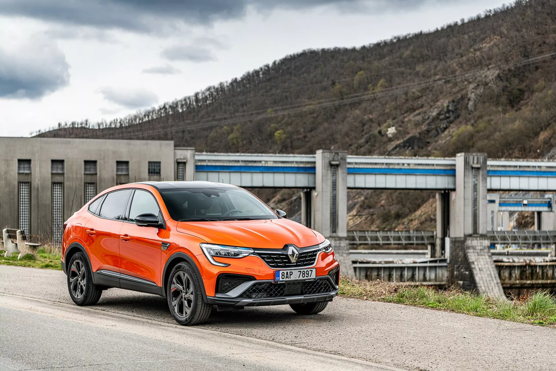 Nowoczesny, jaskrawopomarańczowy crossover SUV Renault Arkana 2021 zaparkowany przy drodze, sfotografowany z perspektywy przednio-bocznej, skupiająca się na stronie kierowcy. Obraz pokazuje przedni grill samochodu z dużym emblematem, eleganckie, kątowe reflektory LED, dzienne światła oraz wloty powietrza i prawdopodobnie czujniki systemów wspomagania kierowcy na dolnym zderzaku, wyraźnie zaznaczone nadkola z wieloramiennymi felgami aluminiowymi i lusterka z zintegrowanymi kierunkowskazami. Pojazd klasyfikowany jako SUV ma podwyższoną postawę z większym prześwitem i pochylony ku tyłowi dach w stylu coupe. W tle widać infrastrukturę przypominającą tamę lub system śluz, co situuje pojazd w przemysłowym lub wiejskim otoczeniu. Ujęcie wykonano z nieco niskiego kąta, co podkreśla wysokość i postawę samochodu.