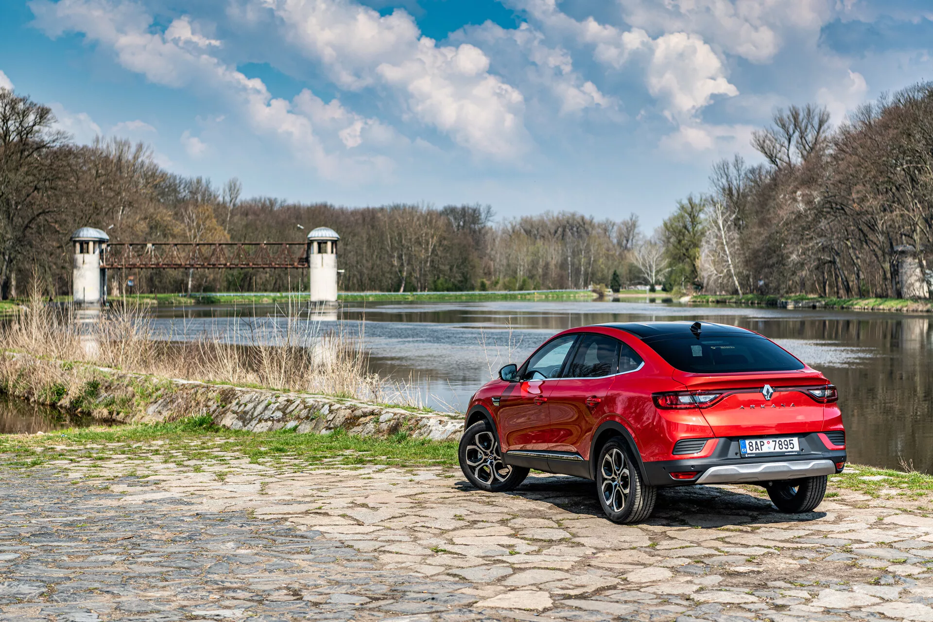 Renault Arkana 2021 zaparkowana na bruku koło wody z naturalnym tłem, prezentująca widok z tyłu z boku, sportową czerwoną konstrukcję i tylne światła LED