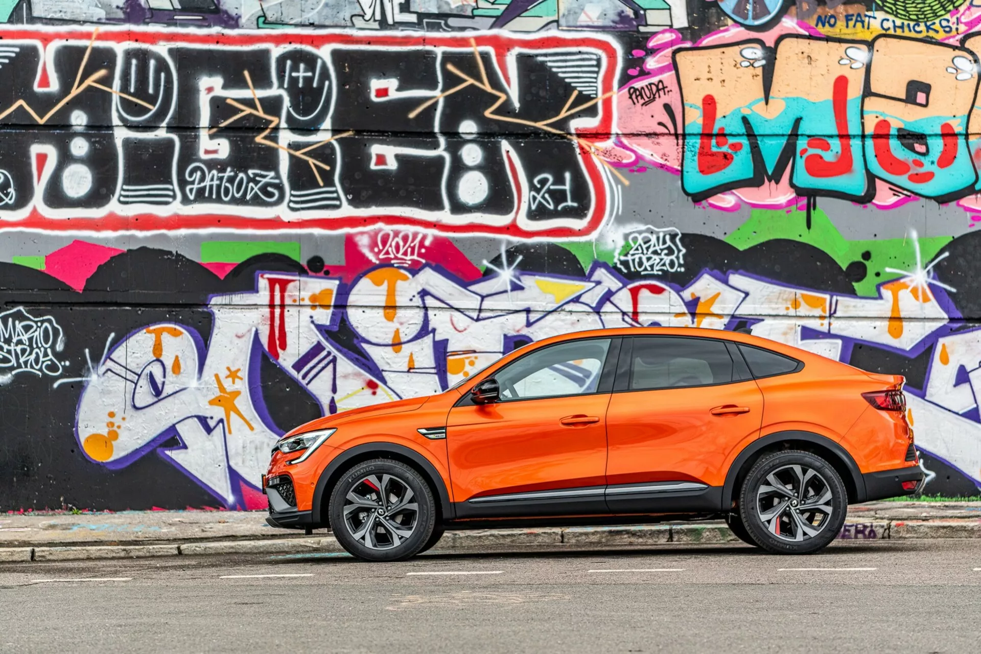 Obraz przedstawia jaskrawopomarańczowy crossover SUV Renault Arkana 2021 zaparkowany równolegle do ulicy z jasno kolorową ścianą graffiti w tle. Zdjęcie uchwycone pod kątem bocznym podkreśla sportowy i nowoczesny design pojazdu z widocznymi przednimi i tylnymi drzwiami kierowcy, bocznymi oknami, lusterkami bocznymi i opadającą linią dachu. Stylizowane felgi aluminiowe z oponami o niskim profilu sugerują skupienie na osiągach drogowych. Klamki bezkluczykowe na przednich drzwiach wskazują na system bezkluczykowego dostępu i uruchamiania. Zdjęcie zostało zrobione w świetle dziennym, podkreślając kontrast pomarańczowego nadwozia na tle graffiti i pokazując dobrze utrzymaną, wyrazistą karoserię w młodzieżowym, miejskim otoczeniu.