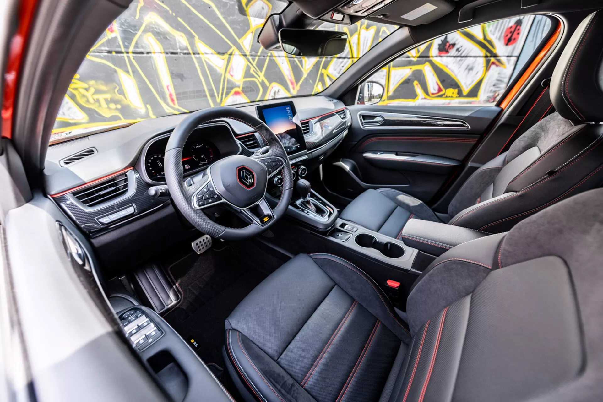 Wnętrze Renault Arkana 2021 prezentujące luksusową przednią kabinę z perspektywy drzwi pasażera z nowoczesną kierownicą trójramienną, dotykowym ekranem systemu infotainment i sportową tapicerką z czerwonymi przeszyciami