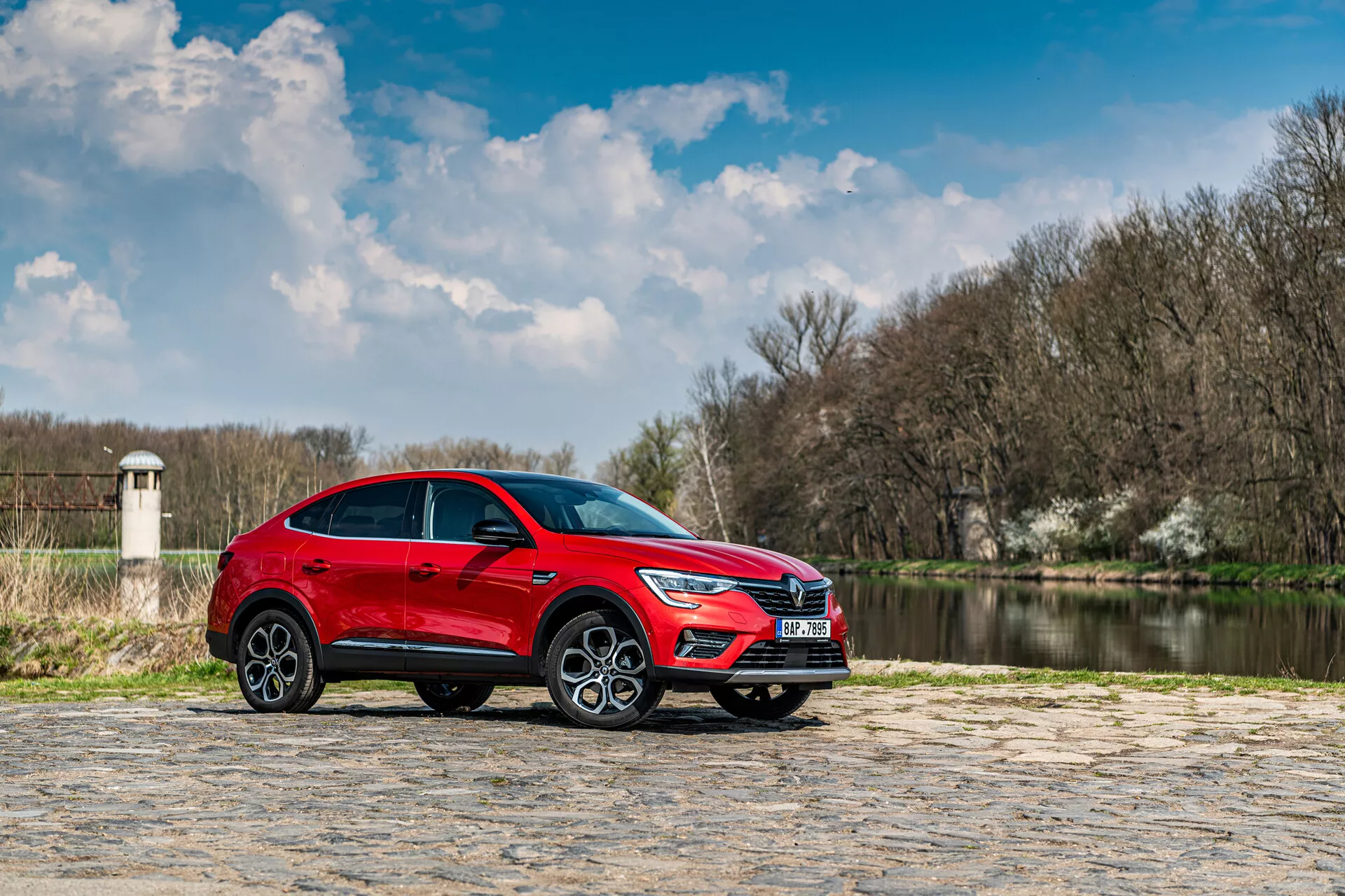 Czerwone SUV Renault Arkana 2021 zaparkowane na kamiennym bruku z wodą i drzewami w tle pod częściowo zachmurzonym niebem, widok z profilu podkreślający elegancki design i dynamiczne funkcje