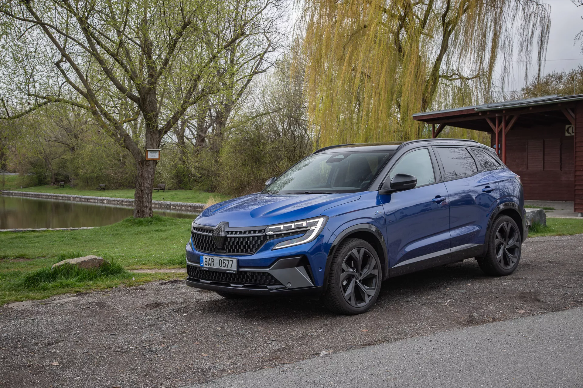 Crossover SUV Renault Austral 2022 w metalicznym, niebieskim kolorze, zaparkowany na zewnątrz pokazujący profil przedni i boczny z reflektorami LED i relingami dachowymi, otoczony naturalnym parkiem