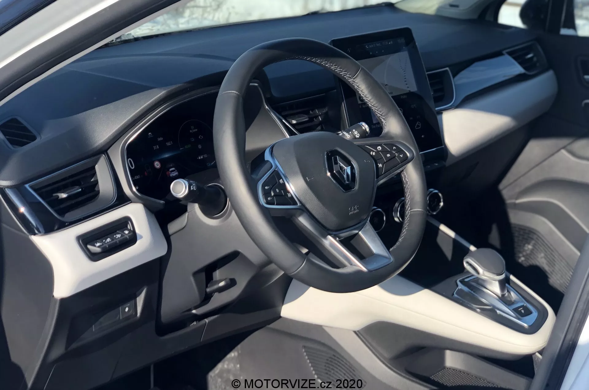 Widok wnętrza Renault Captur 2020 z perspektywy kierowcy, ukazujący kierownicę z logo Renault, zamontowane przyciski, cyfrowy zestaw wskaźników z tyłu i centralnie umieszczony ekran dotykowy systemu informacyjno-rozrywkowego. Zdjęcie podkreśla mieszankę materiałów syntetycznych i plastikowych z metalicznymi akcentami, automatyczną skrzynię biegów, system kontroli klimatu i regulację szyb i luster.