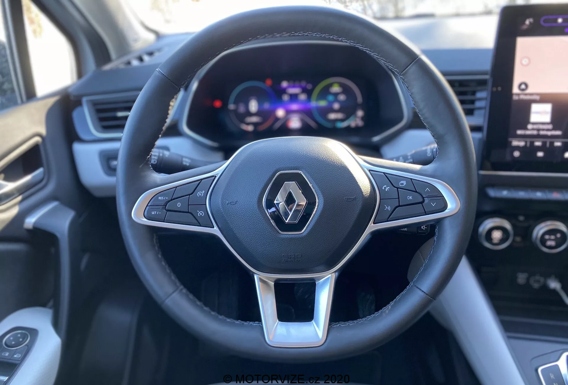 Zbliżenie z perspektywy pierwszej osoby z fotela kierowcy Renault Captur 2020, skupiający się na wielofunkcyjnej kierownicy trójramiennej obszytej skórą z srebrnym akcentem i logo Renault, w towarzystwie kolorowego cyfrowego zestawu wskaźników wyświetlającego prędkościomierz i szczegóły pojazdu, z częściowo widocznym ekranem dotykowym infotainmentu w tle.