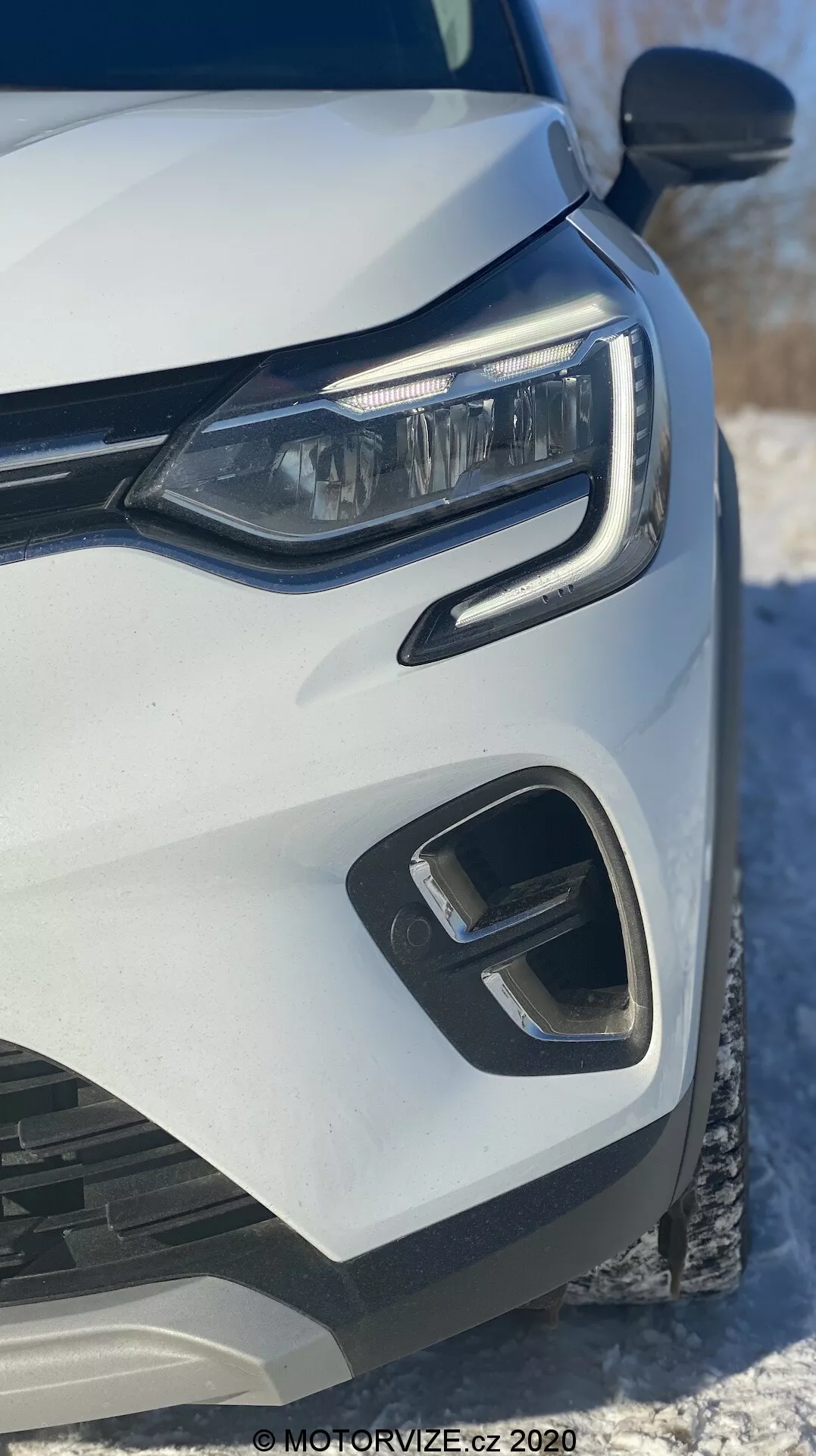Zbliżenie na przednią lewą stronę białego Renault Captur 2020, z naciskiem na nowoczesny reflektor LED, geometryczny przedni zderzak i elementy kratki, uchwycony pod niskim kątem czołowym, z widocznym śniegiem w tle