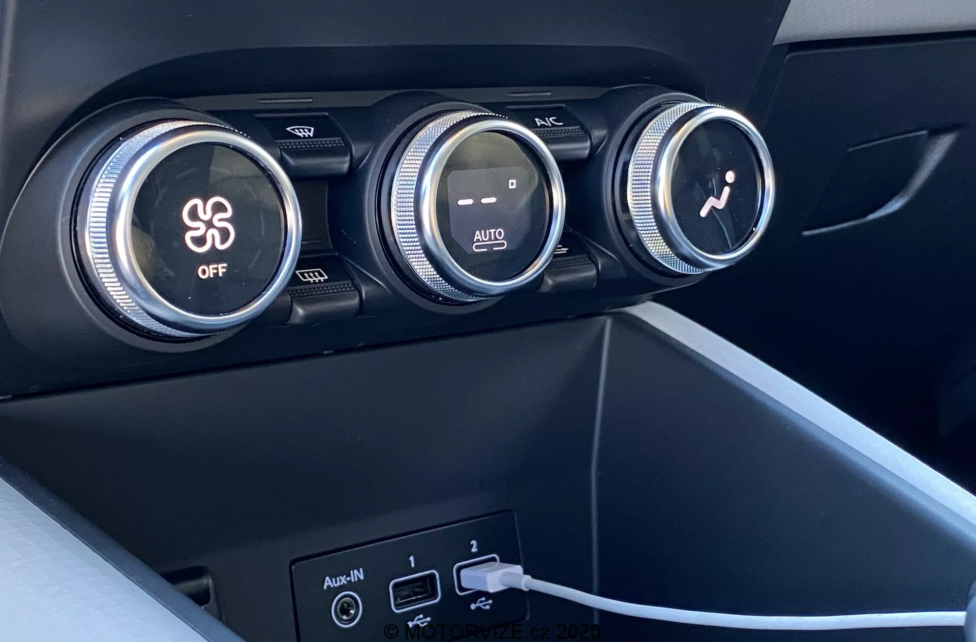 Zbliżenie wnętrza Renault Captur 2020 skupiające się na systemie klimatyzacji. Na zdjęciu widać trzy duże pokrętła na desce rozdzielczej. Pierwsze pokrętło kontroluje klimatyzację, wskazane symbolem płatka śniegu i przyciskiem 'OFF'. Środkowe pokrętło reguluje temperaturę za pomocą czerwonych i niebieskich gradiacji kolorów oraz oznaczenia 'AUTO'. Trzecie pokrętło z ikoną wentylatora reguluje prędkość wentylatora. Poniżej pokręteł znajdują się dwa porty USB i wejście pomocnicze, z podłączonym kablem do jednego z portów USB. Materiały to ciemny plastik lub tworzywo sztuczne ze szlifem matowym oraz nowoczesny, minimalistyczny design.