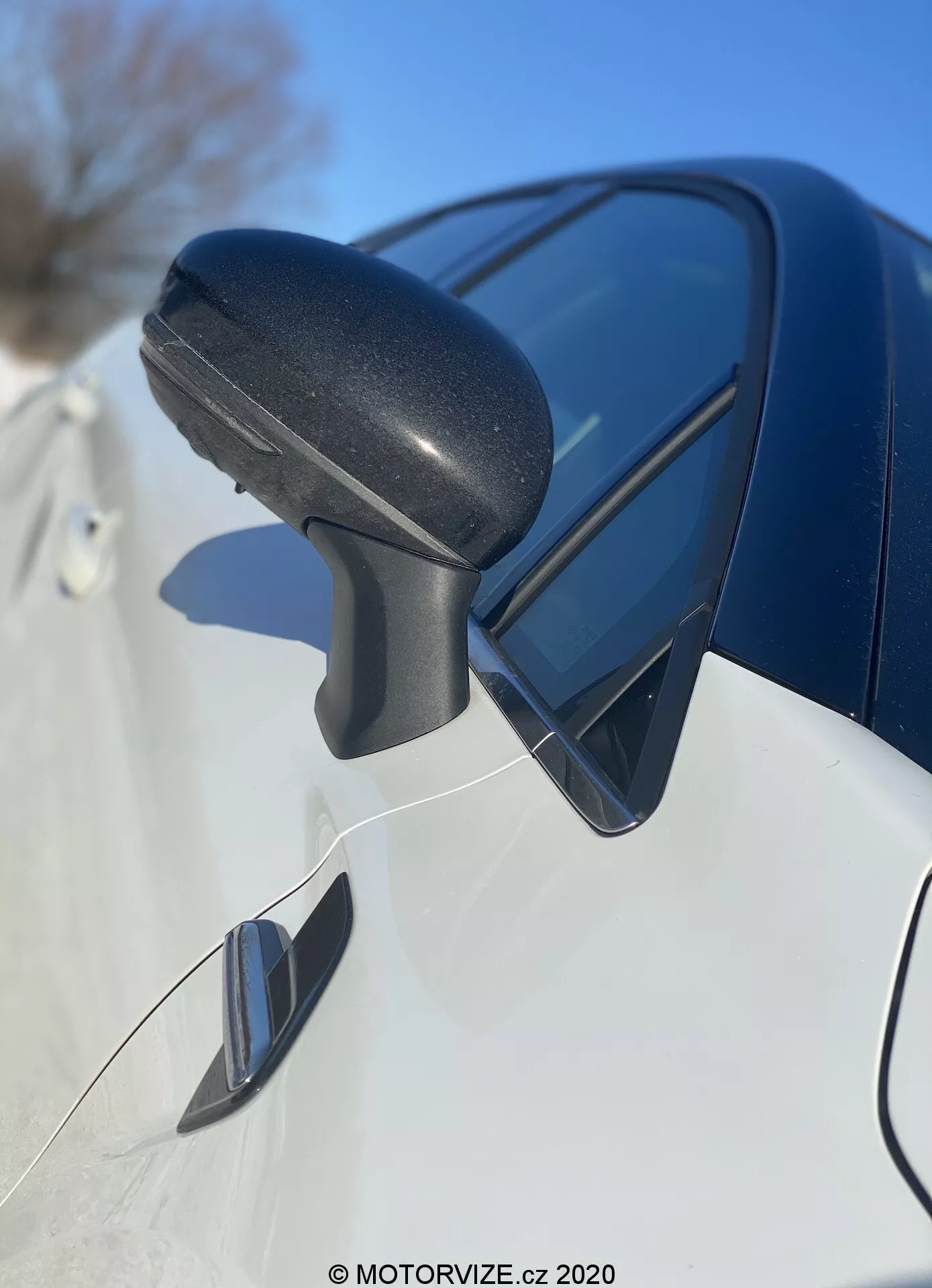 Zbliżenie obudowy lusterka bocznego Renault Captur 2020 od przodu do tyłu, z naciskiem na czarną teksturowaną plastikową obudowę lusterka z wbudowanym sygnalizacją skrętu i odbijającym niebo. Elegancki ciemny panel karoserii poniżej i potencjalne lusterko punktu ślepego wskazują na nowoczesny design i zaawansowane funkcje.