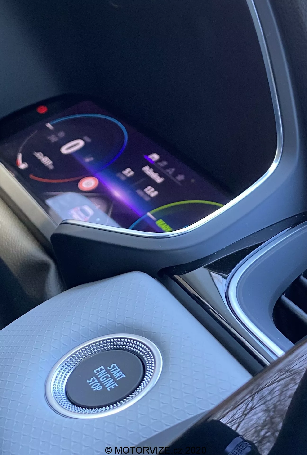 Wnętrze Renault Captur 2020 z naciskiem na konsolę środkową i deskę rozdzielczą z perspektywy siedzenia przedniego. Żywy ekran infotainment wyświetla kolorowy interfejs sugerujący zaawansowane funkcje multimedialne, w tym nawigację, odtwarzanie multimediów i opcje łączności. Poniżej ekranu znajduje się okrągły przycisk sterowania z chromowanym wykończeniem oznaczony 'Engine Start Stop', co podkreśla system bezkluczykowego uruchamiania. Konsola środkowa ma teksturowane wykończenie wysokiej jakości, co poprawia atmosferę wnętrza. Po lewej stronie częściowo widoczny jest uchwyt na kubek lub schowek.